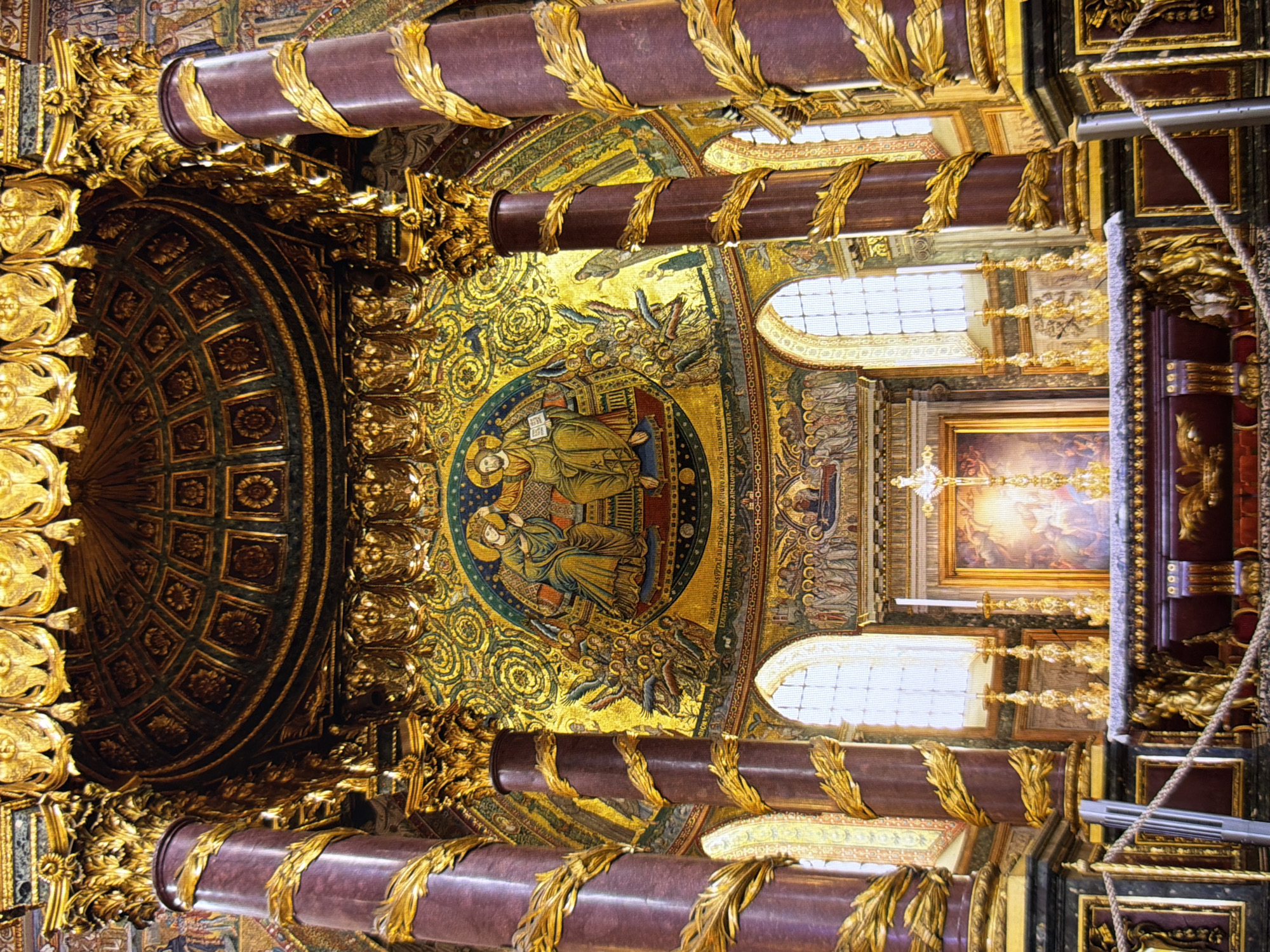 This image depicts the interior of a richly decorated church or cathedral. Prominently featured are two large, dark red columns adorned with ornate gold leaf detailing that spirals upwards. Above, a domed ceiling, also intricately decorated with gold and dark green patterns, is visible.
The focal point of the scene is a large, gilded mosaic on the back wall. This mosaic features depictions of religious figures, including what appear to be Jesus Christ and possibly the Virgin Mary, surrounded by a multitude of smaller figures and angelic beings. Below the mosaic is a framed painting depicting a religious scene, with a cross and candles placed in front of it on an altar.
The altar itself is adorned with decorative elements, including what seem to be figures and gold leaf. In the foreground, a rope barrier with metal posts is visible, suggesting a roped-off area for visitors. Large arched windows with patterned glass are positioned behind the altar, admitting natural light. The overall impression is one of opulence, with extensive use of gold, mosaics, and intricate carvings, indicative of a significant religious site.
The scene described is not located in Kyiv, Ukraine, but rather appears to be an interior of a historic religious building in Rome, Italy, likely the Basilica di Santa Maria Maggiore, given the distinctive apse mosaic.