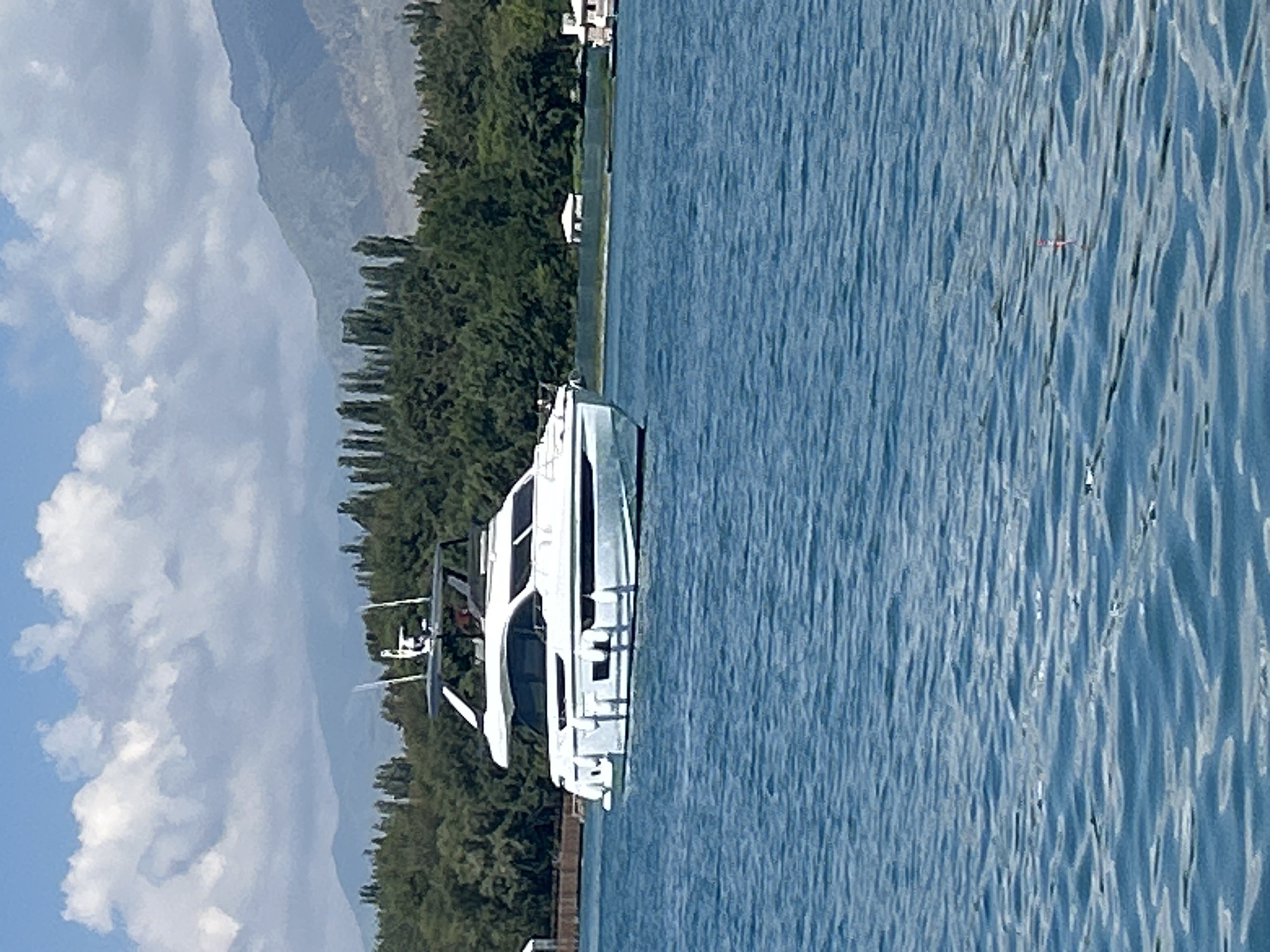 A white luxury yacht is positioned on the blue waters of Issyk-Kul Lake in Cholpon-Ata, Kyrgyzstan. The yacht has a sleek, modern design with dark tinted windows and a white radar arch. In the background, a dense line of green trees separates the lake from rolling hills and mountains. The sky is a clear blue with scattered white clouds. A small red and white fishing float is visible on the water's surface in the foreground.