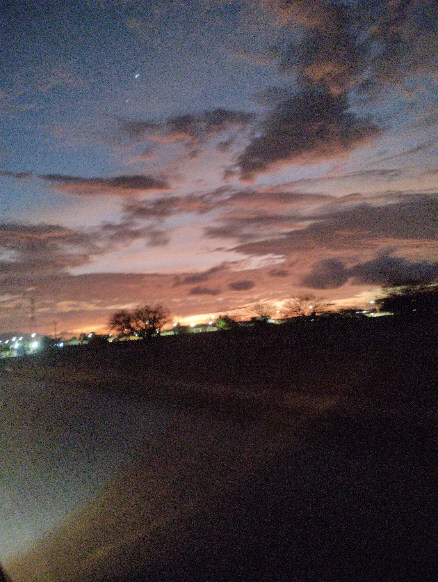 This image captures a twilight sky over a somewhat rural or suburban area, possibly on the outskirts of El Espinal, Venezuela. The sky is a blend of deep blues and purples, transitioning into warmer orange and pink hues as they reflect the setting sun. Wispy clouds are scattered across the expanse, with some appearing darker and more defined than others.
In the middle ground, a silhouette of trees is visible, their branches bare against the colorful sky, suggesting a drier season. Beyond the trees, a line of lights indicates a settlement or town. These lights are small and somewhat diffused, consistent with streetlights or distant building illumination. A power line tower is also faintly visible on the left side of the image.
The foreground is dominated by a dark, textured surface, likely a road or unpaved ground. The overall impression is one of a quiet evening, with the natural beauty of the sky contrasting with the subtle signs of human presence in the distance. There are no people or specific identifiable objects in the foreground or mid-ground.