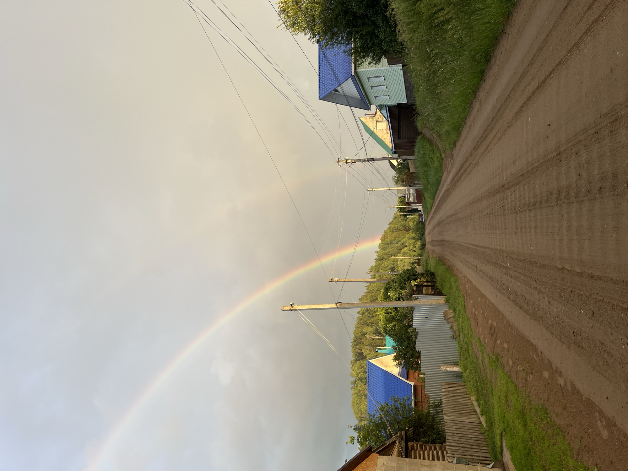 This image depicts a rural village road in the village of Olkhovka, Russia, under a sky with a double rainbow. The road is unpaved and has visible tire tracks. On the left side of the road, there are several wooden houses with fences and some greenery. On the right side, there are more houses, some with blue roofs, and abundant vegetation. Telephone poles with wires are present along the road. The overall impression is that of a peaceful, natural setting, possibly after a rain shower.
