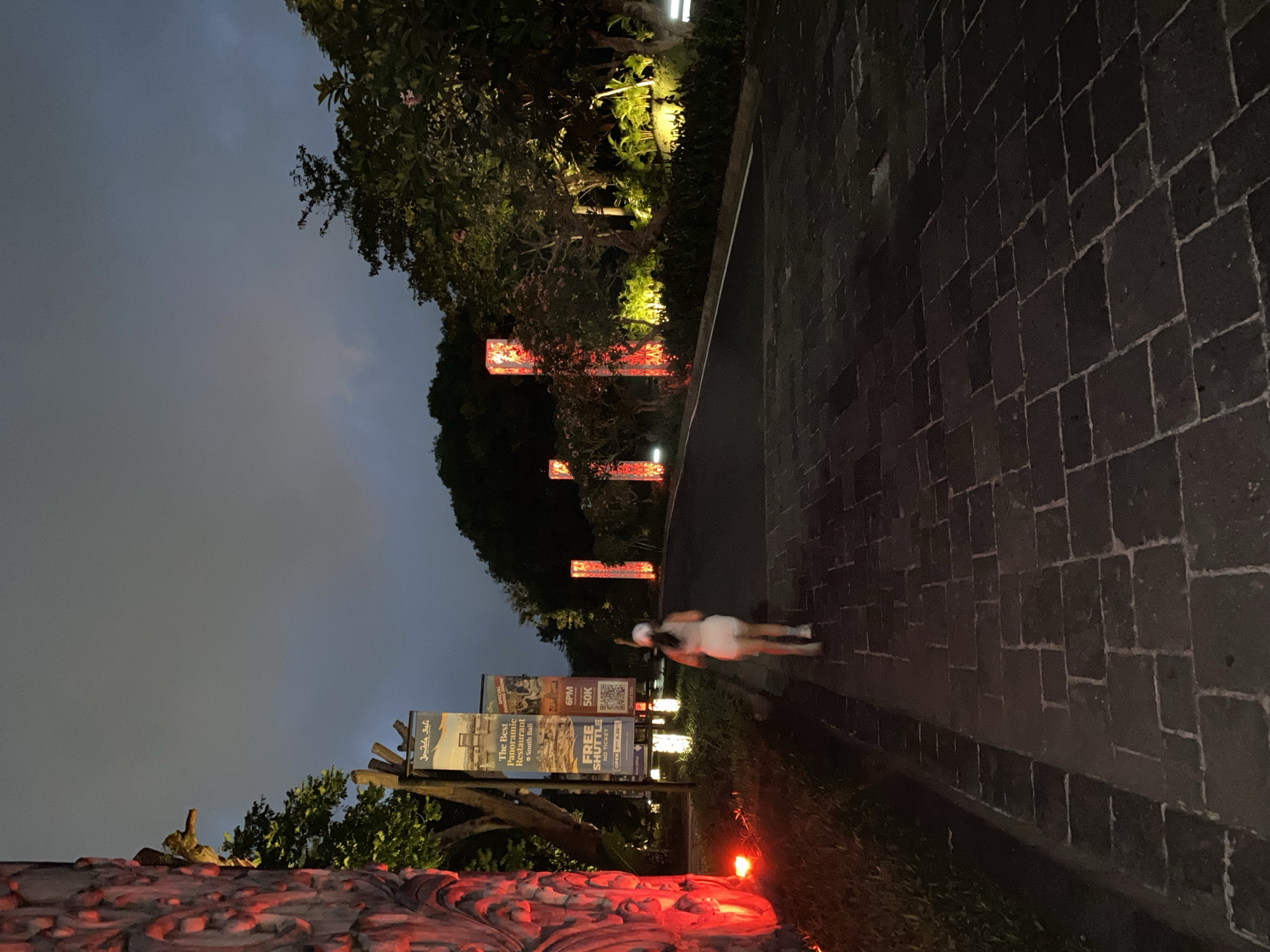 In Kuta Selatan, Indonesia, a solitary person walks away from the camera down a paved pathway at dusk. The pathway is lined with dark, rectangular stones and illuminated by intermittent red and white lights. To the left, a large, intricately carved stone pillar glows with red light, and a banner displays text including "The Best Panoramic Restaurant" and "South Bali." On the right, trees and foliage are lit from below, creating a warm ambiance. The sky is overcast, and a few tall, red-lit structures stand as decorative elements along the pathway.