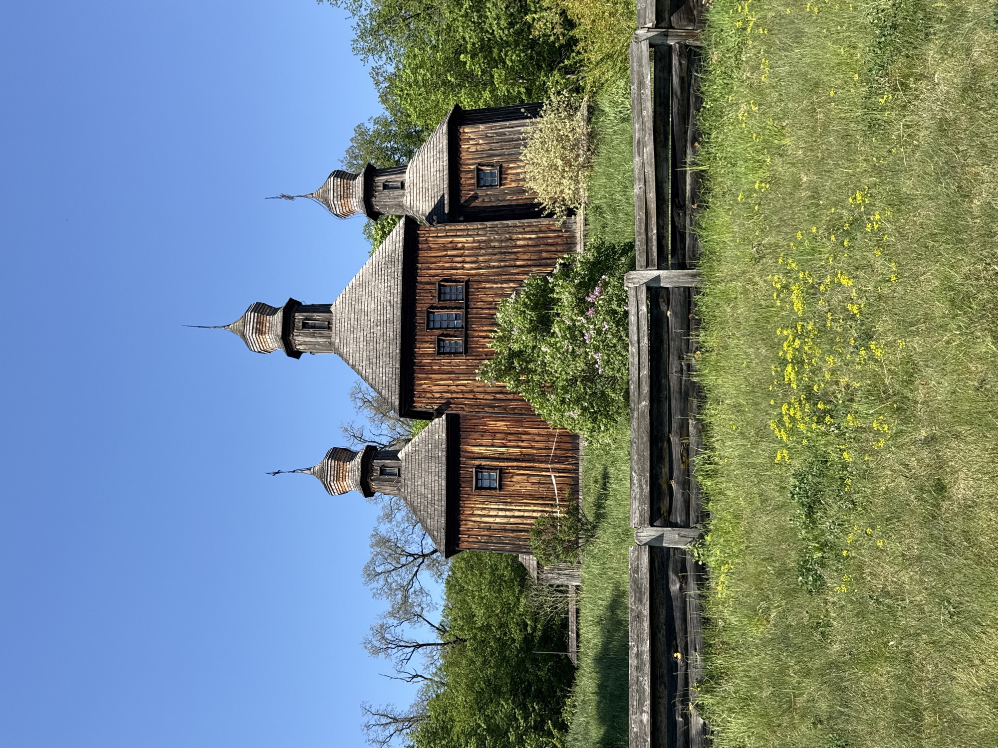 The image captures an old wooden church in Kyiv, Ukraine. The church, constructed from dark wood, features a multi-tiered structure with distinct steeples capped with a conical design. The roof appears to be made of shingles. Small windows punctuate the facade. A rustic wooden fence runs alongside a field of tall green grass and yellow wildflowers. Trees surround the church, adding to the natural setting. The sky is clear and blue.