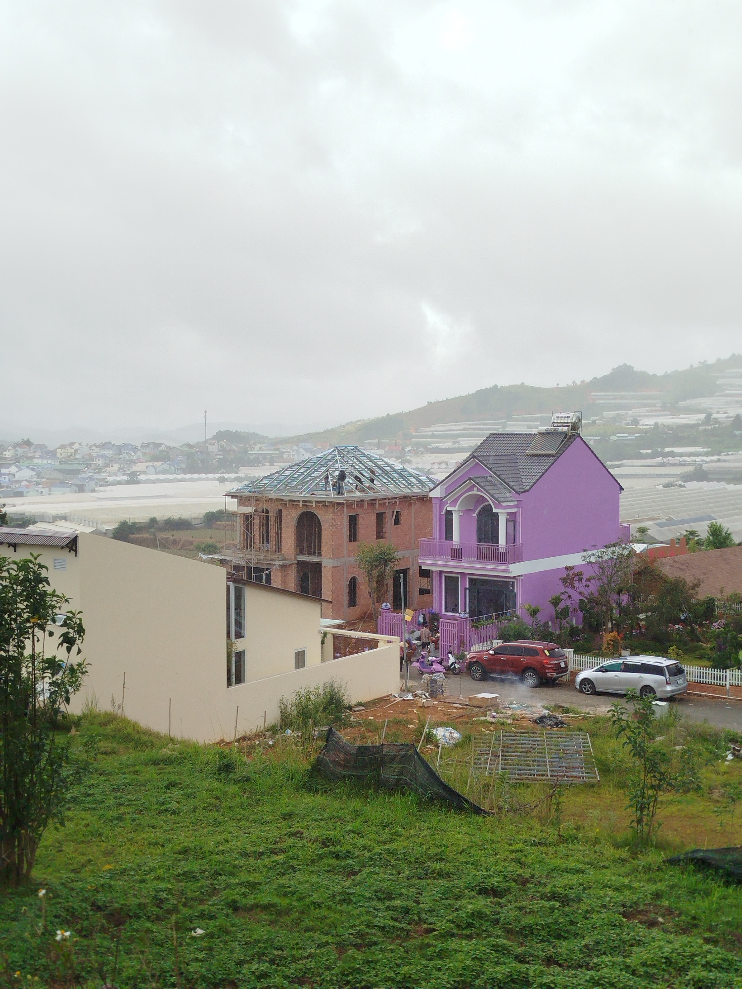 This image depicts a residential construction site in Lạc Dương, Vietnam, under a cloudy sky. In the foreground, a grassy slope is visible with some scattered debris and construction materials, including a metal frame and black netting.
Two houses are prominently featured. To the left, a beige, two-story house with modern architecture is partially visible. To its right, a brick house under construction shows its framework for the roof, with at least two individuals working on it. Further to the right, a vibrant purple, multi-story house stands completed, featuring a balcony and a solar water heater on its roof.
In front of the purple house, a red SUV and a silver station wagon are parked. A white picket fence lines the property of the purple house. In the background, the landscape transitions to a town with numerous buildings, and beyond that, rolling hills covered with greenhouses, indicating agricultural activity. The overall atmosphere is overcast and slightly hazy.
