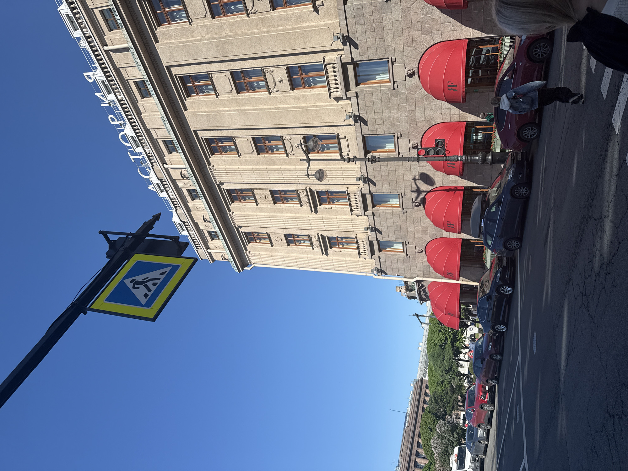 An urban scene in Saint Petersburg, Russia, is captured on a clear, sunny day. The dominant feature is a grand, multi-story building constructed from light-colored stone, featuring numerous rectangular windows with brown frames and intricate architectural details. Along the ground floor, several vibrant red, semicircular awnings project outwards, each displaying the white initials "BF." High on the building's facade, a large sign with white Cyrillic letters spells "АСТОРИЯ" (ASTORIA), identifying the establishment as the Astoria.
Adjacent to the building is a paved street with white markings, where a row of cars, including red and dark-colored sedans, are parked along the curb. A black traffic light pole with a red signal is visible near the building. A person wearing a light denim jacket and dark pants is seen walking on the sidewalk next to the parked cars, moving away from the camera. Other individuals are faintly visible further down the street.
In the foreground, a yellow-bordered blue pedestrian crossing sign, featuring a white walking figure, is mounted on a black pole, set against a bright, clear blue sky. The strong sunlight and cloudless sky indicate a pleasant daytime setting. The scene portrays typical urban activity, with pedestrians and parked vehicles contributing to the city atmosphere.