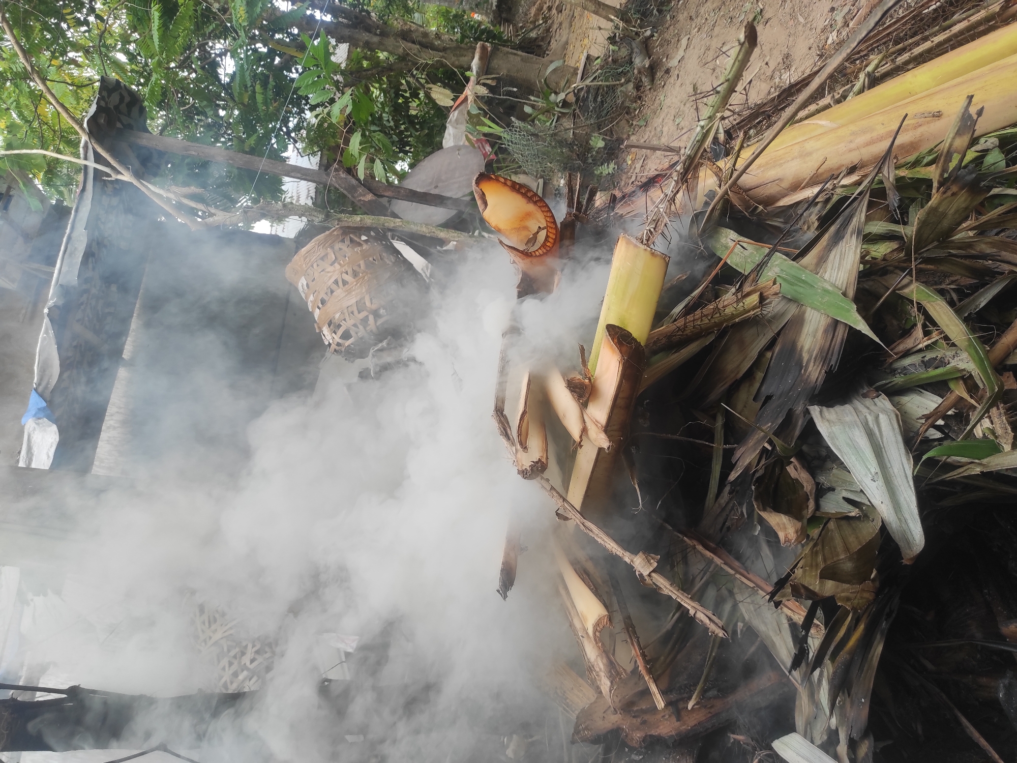 A pile of dried plant matter, likely banana tree remnants and leaves, is burning and producing a significant amount of smoke. The scene appears to be an outdoor area, possibly a backyard or open lot, with trees and vegetation visible in the background. A makeshift shelter with a corrugated metal roof is partially visible behind the smoke. The ground is dirt and appears somewhat uneven. There are no people or animals clearly visible in the image. The overall impression is of a controlled burn or the disposal of organic waste. The location is identified as Lhokseumawe, Indonesia.