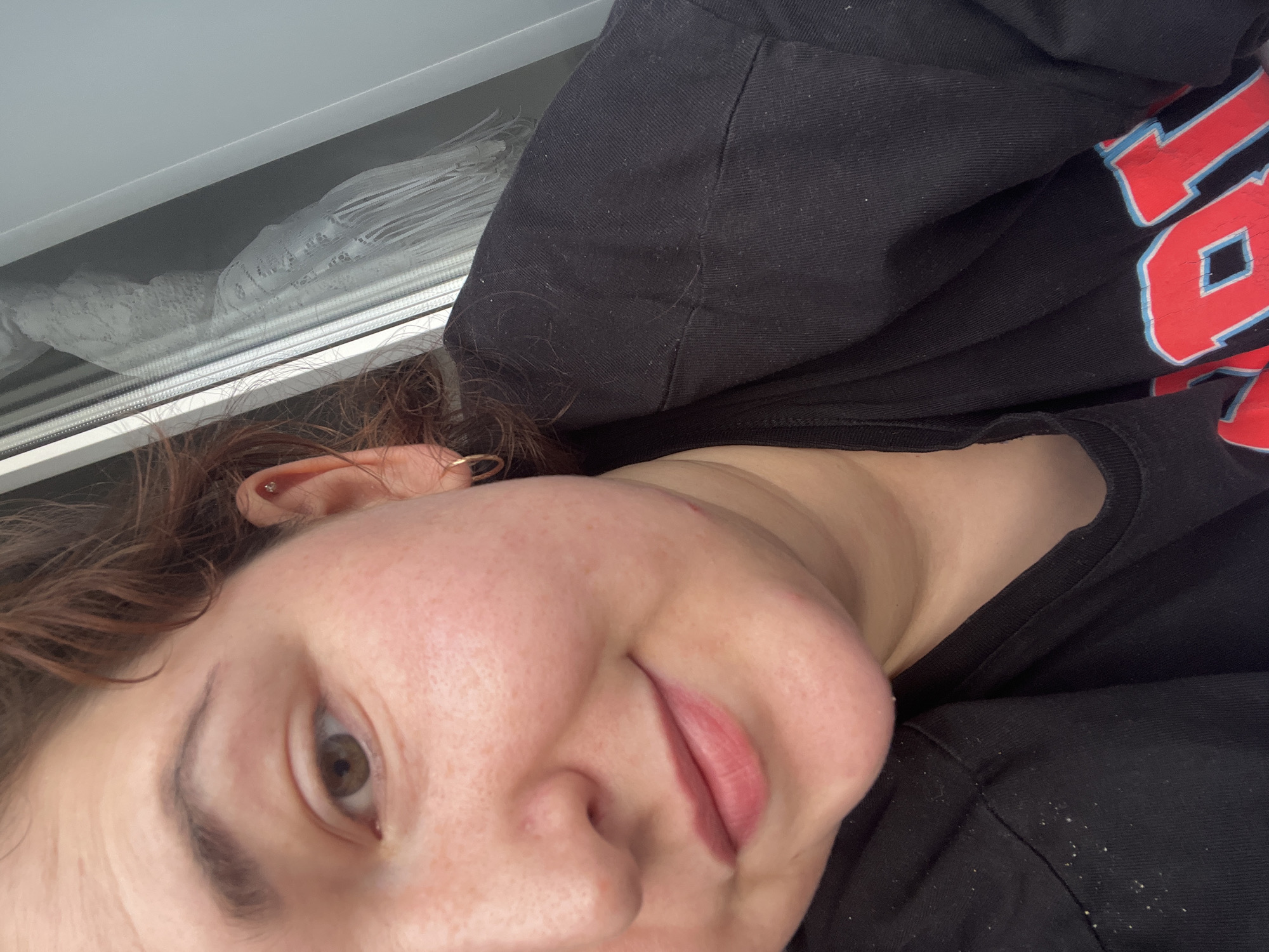 The image is a close-up, upper-body portrait of a person with fair skin, reddish-brown hair, and brown eyes. The person is wearing a black t-shirt with red and blue lettering. They have a small hoop earring in their left ear and a stud earring in their upper left ear. The background shows a window with white curtains and a grey frame. The person appears to be indoors, with natural light coming from the window. There is no identifiable location in the city of Chelyabinsk, Russia, visible in the image.