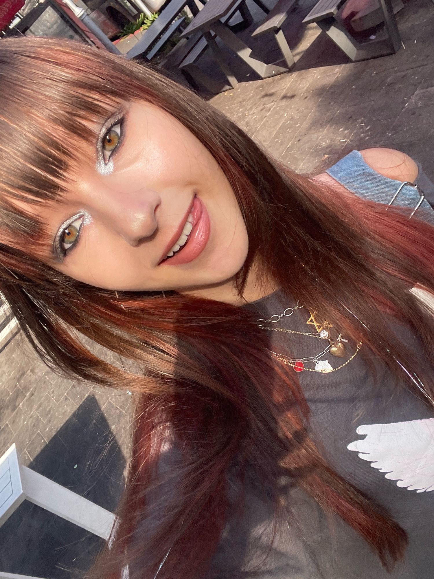 A young woman with auburn hair, featuring prominent bangs and streaks of red, is captured in a close-up selfie. She is outdoors, possibly at a cafe or restaurant with tables and chairs visible in the background, with sunlight casting shadows on the paved ground. The woman is wearing a dark t-shirt with a white graphic design of a bird's wing and a multi-layered necklace adorned with charms including a Star of David, a heart, and enamel pendants. Her makeup includes shimmering silver eyeshadow and glossy pink lipstick, and she is sticking her tongue out slightly, giving a playful expression. The background suggests an urban setting, consistent with Raanana, Israel.