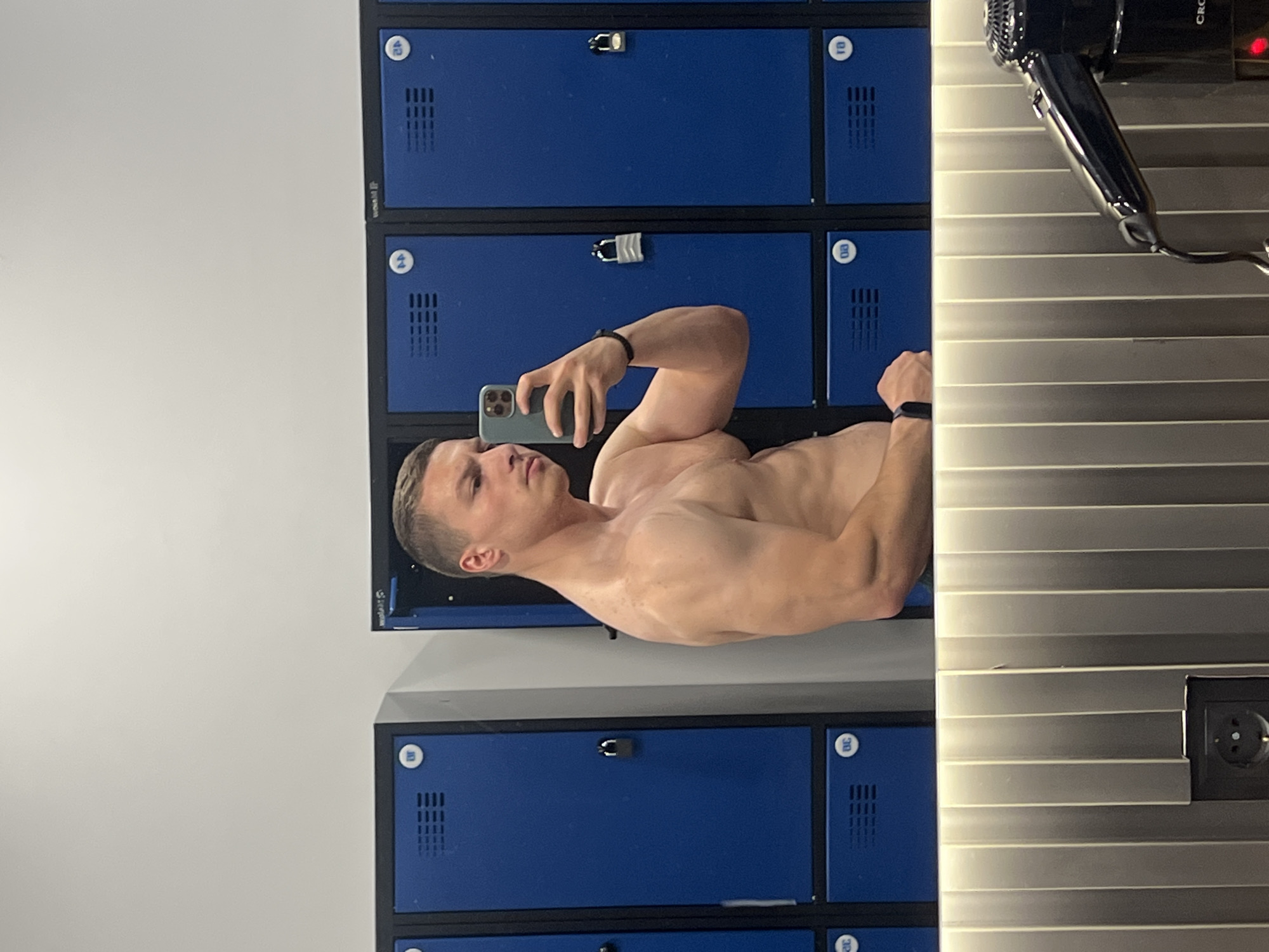 A shirtless, muscular man is taking a selfie in what appears to be a gym locker room. He is holding his phone with his right hand, flexing his left arm, and standing in front of a wall of blue lockers. The background also includes a gray, horizontally striped wall with a hair dryer attached. An electrical outlet is visible on the wall. The location is plausibly Burgas, Bulgaria, given the presence of lockers reminiscent of those found in gyms and public facilities in the region.