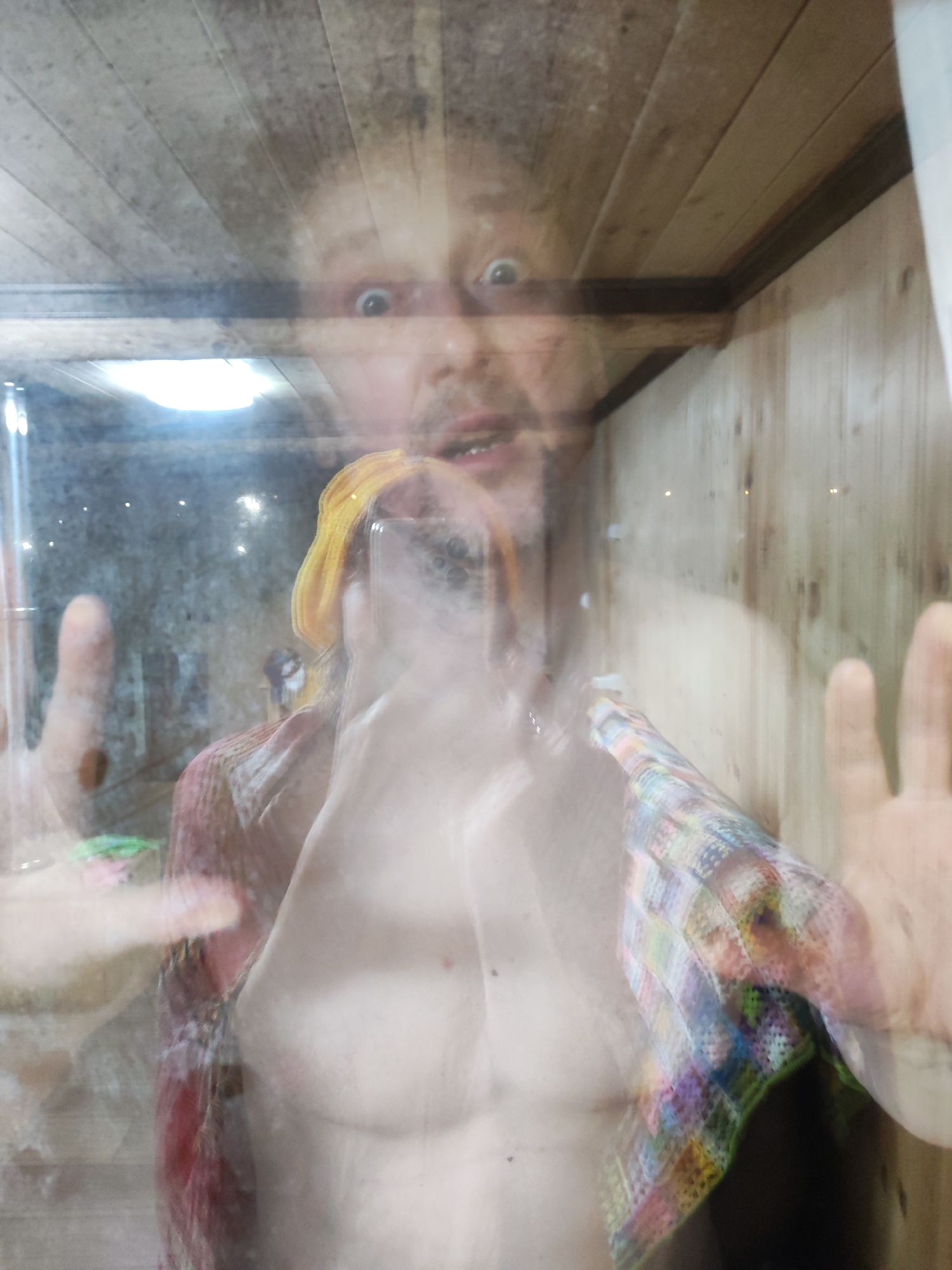 A selfie of a topless man pressed against glass is overlaid with a blurred interior scene. The man's face is visible near the top center of the frame with wide eyes and a slightly open mouth. The blurred interior scene appears to show a room with wood paneling on the ceiling and walls. The man is holding a phone in front of him, partially obscuring his torso. The location is Chaikovsky, Russia.