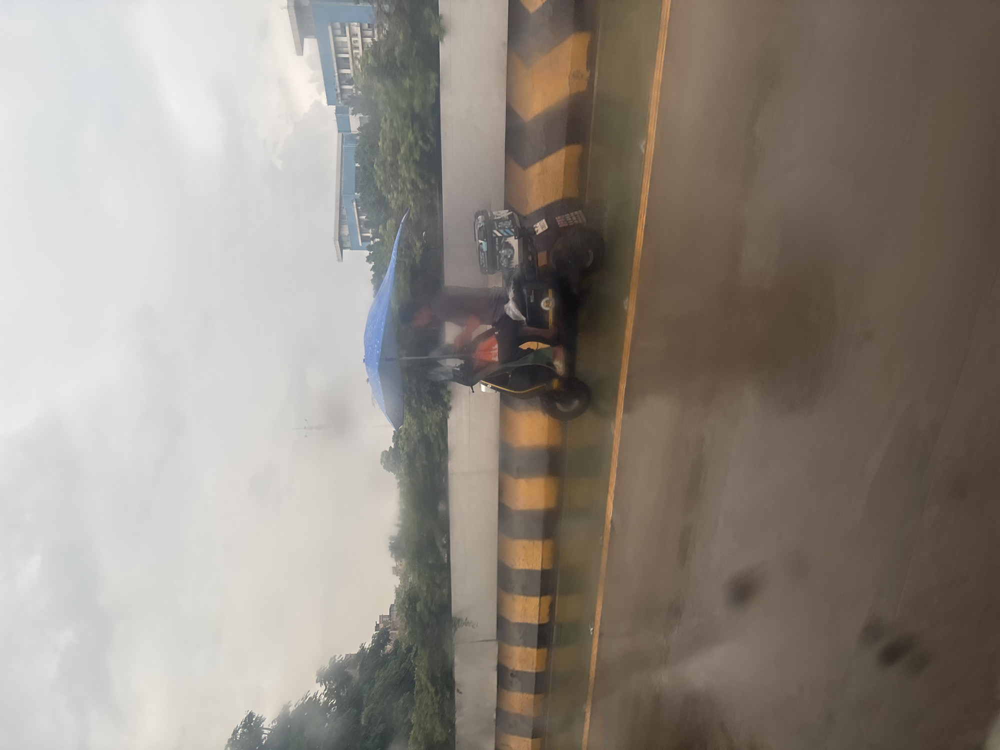 On a rain-slicked road in Nanhai District, China, two individuals share a scooter, shielded by a bright blue umbrella. The scooter is dark-colored with a black storage box affixed to the rear. A concrete barrier with alternating black and yellow stripes runs parallel to the road, separating it from a grassy embankment topped with lush green trees and buildings. The sky is overcast and a hazy gray, suggesting persistent rainfall. The scene captures a moment of everyday life on the move amidst inclement weather.