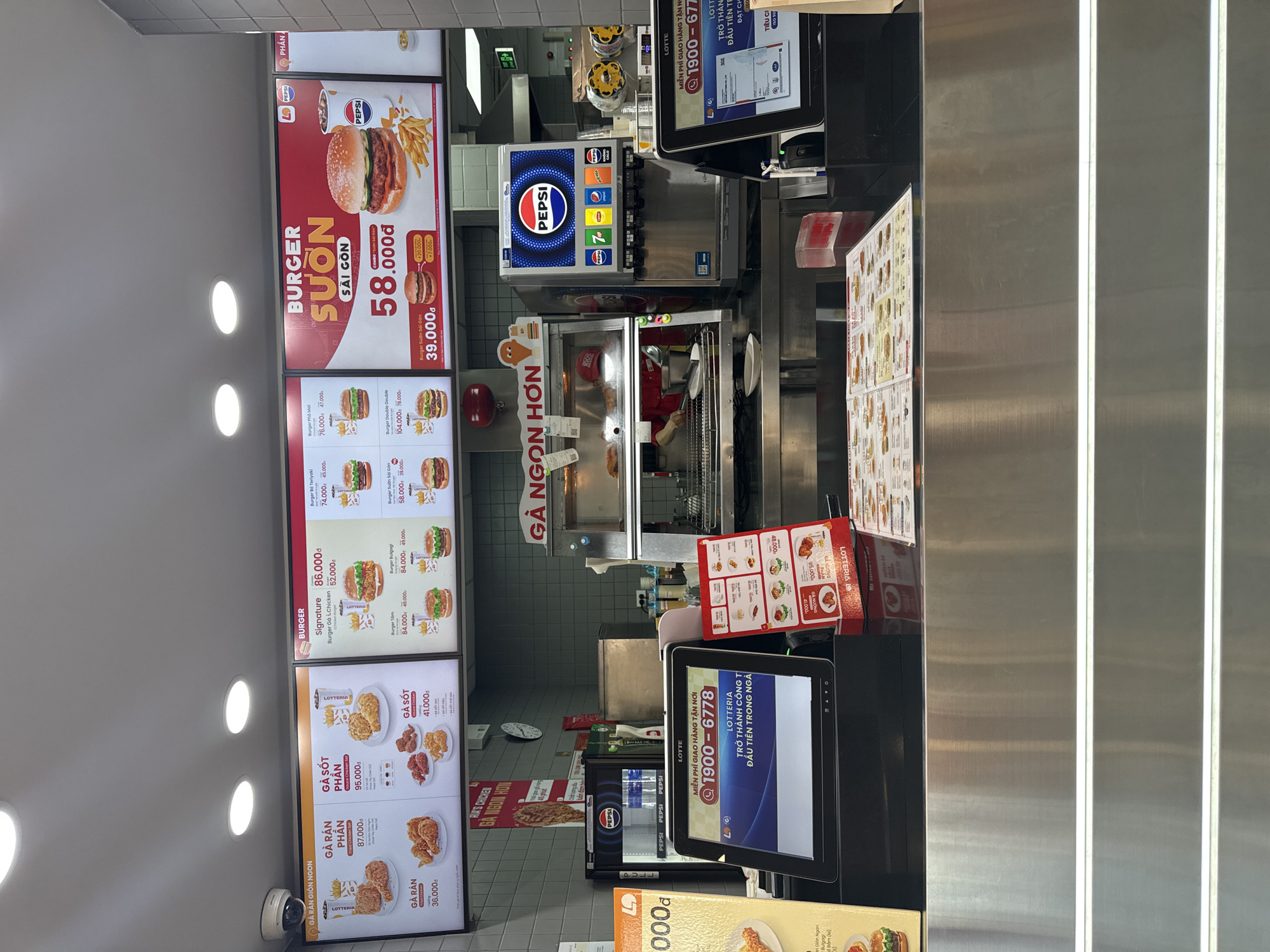 The image shows the interior of a Lotte Chicken fast-food restaurant, likely located in Bac Nha Trang Ward, Vietnam, based on the context of typical Lotte Chicken locations. The scene is dominated by a brightly lit menu board displaying various chicken and burger options. In the foreground, a Lotte-branded touchscreen ordering system is visible, along with a promotional stand featuring menu items. Behind the counter, a Pepsi beverage dispenser is prominent, with several employees in uniform visible working in the kitchen area. The overall environment appears to be a standard fast-food service counter, with clean stainless steel surfaces and food preparation equipment. The image captures a typical operational moment within a fast-food establishment.