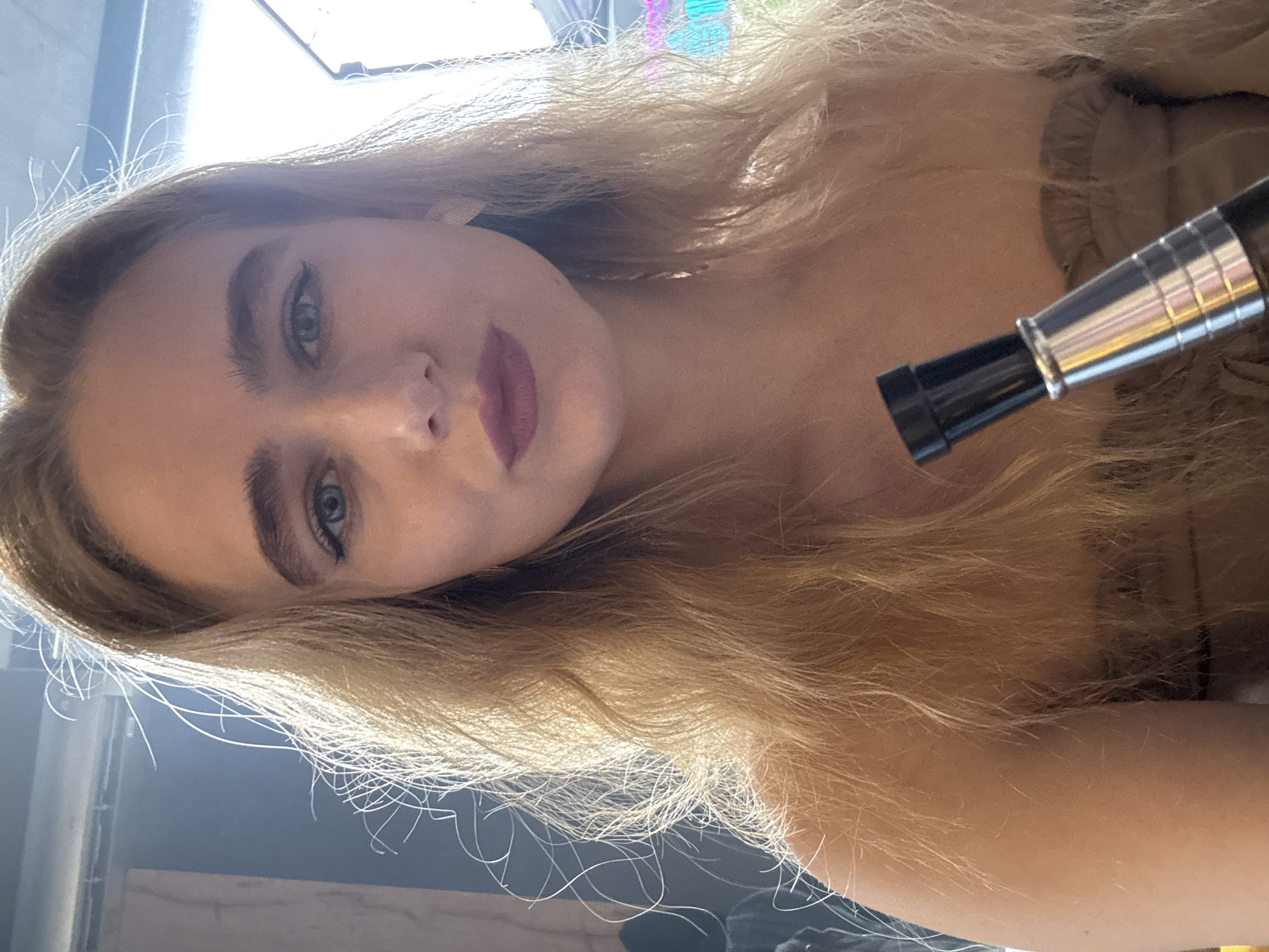 A close-up, tilted, indoor shot features a fair-skinned woman with long, light-blonde hair. She has blue eyes and wears lipstick. A metallic and black object, possibly a cosmetic applicator, is positioned near her right shoulder. The background includes a blurred, illuminated structure and a greyish surface. The location and city are not discernible.