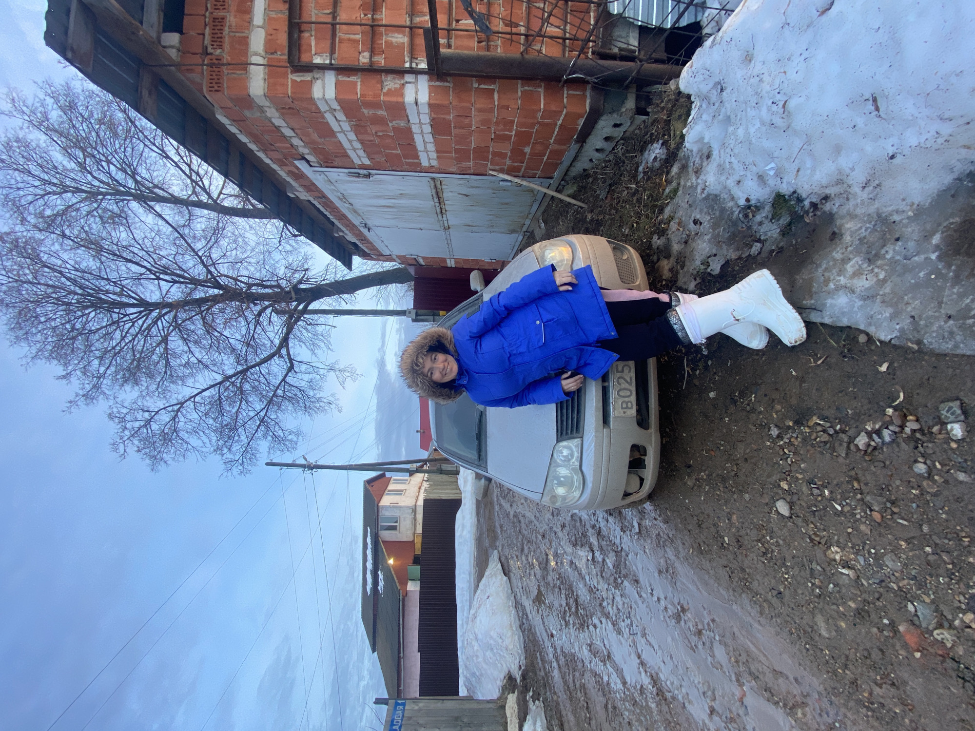 The image displays an outdoor scene on an unpaved, muddy street in Ольховка, Russia, identified by a wooden signpost reading "АЛЛЕЯ 1" (Alley 1). The environment suggests late winter or early spring conditions, with bare deciduous trees, significant patches of melting dirty snow, and a pale blue, partly cloudy sky indicative of twilight.
A female subject, smiling and facing the camera, stands with her left hip leaning against the front-left fender of a light-colored sedan. She is wearing a blue winter parka with a fur-trimmed hood, black pants with a pink stripe along the outer seam, and white winter boots. The sedan's front bumper on the driver's side is visibly damaged, and a partial license plate "B025" is affixed to the front.
To the subject's right, a red brick garage or outbuilding with horizontal white mortar lines features a weathered, light-colored metal double-door and a corrugated metal roof overhang. A bare tree trunk rises immediately behind this structure. A wooden utility pole with multiple overhead wires is visible to the left of the garage and above the car. In the background, to the left of the scene, a two-story house with a reddish-orange lower level and a light-colored upper level stands behind a dark corrugated metal fence; its roof shows patches of snow. The ground is characterized by wet, dark mud mixed with gravel, puddles, and irregular mounds of melting, discolored snow.