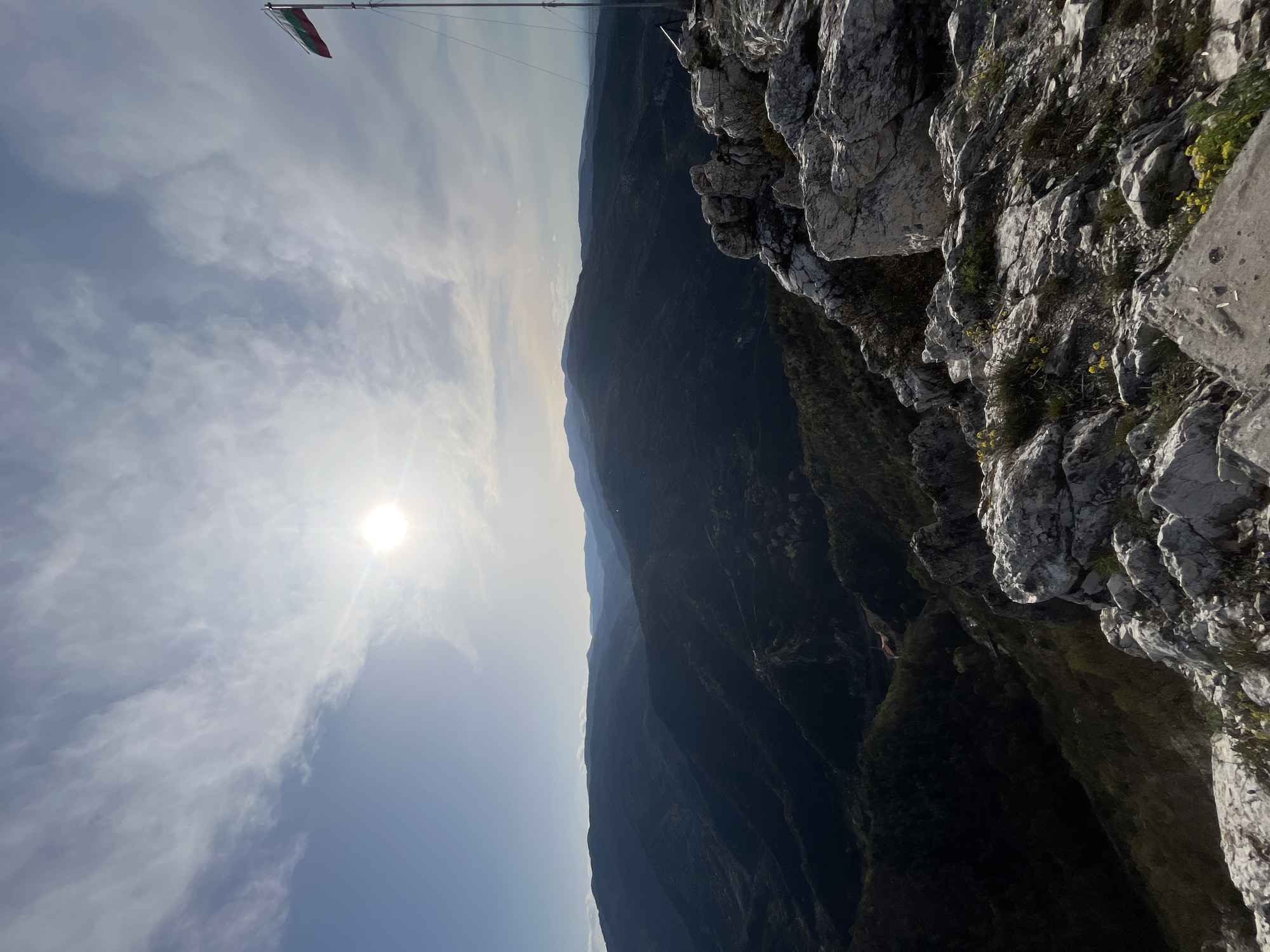 The image shows a mountainous landscape with a rocky cliff in the foreground. The sun is visible in the sky, partially obscured by clouds. A flag, possibly the Bulgarian flag, is mounted on a pole at the top of the cliff. The location is potentially near Asenovgrad, Bulgaria, given the landscape characteristics. The scene is observed from a high vantage point.