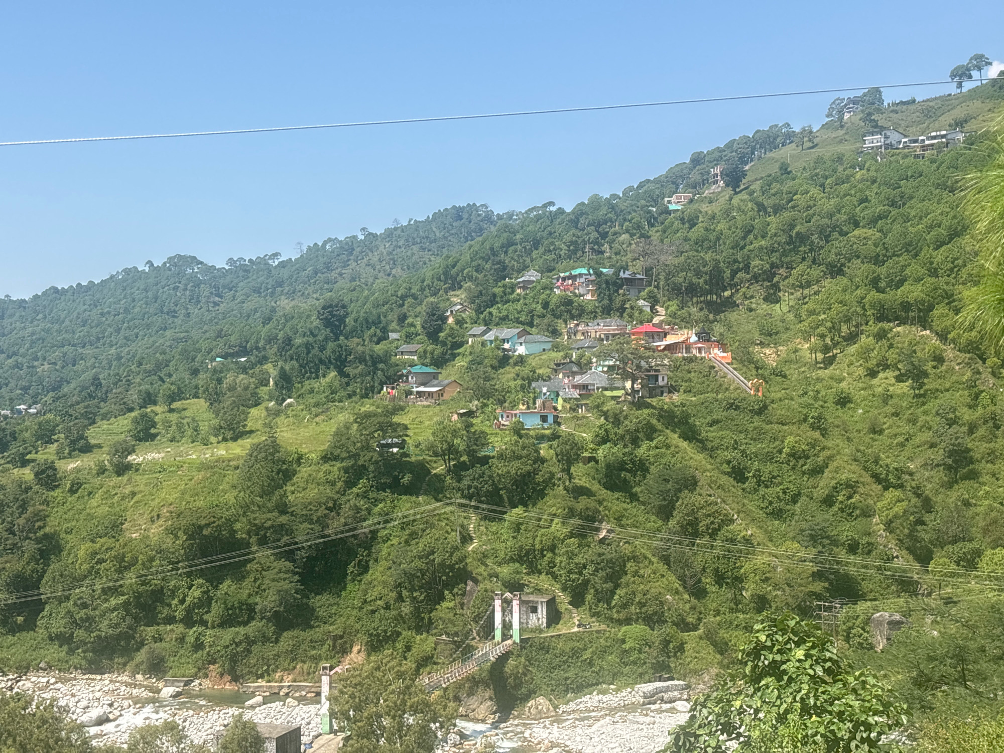 This is a wide-angle, eye-level, outdoor, daytime, horizontal shot of a lush green mountainside village in Palampur, India. The scene is dominated by rolling hills covered in dense, verdant vegetation and scattered trees. Nestled into the slopes are numerous houses, some with red and green roofs, clustered together in small settlements. A riverbed with a rocky, dry appearance runs along the bottom of the frame, with a small, rustic suspension bridge spanning it. The bridge appears to be made of wood and metal, with supporting pillars adorned with green and white paint. Above the mountainside, a clear, pale blue sky stretches across the horizon. Power lines are visible in the upper portion of the image, crossing the sky and traversing the mountainside. No people are visible in the frame. The overall impression is one of a peaceful, rural, mountainous landscape.