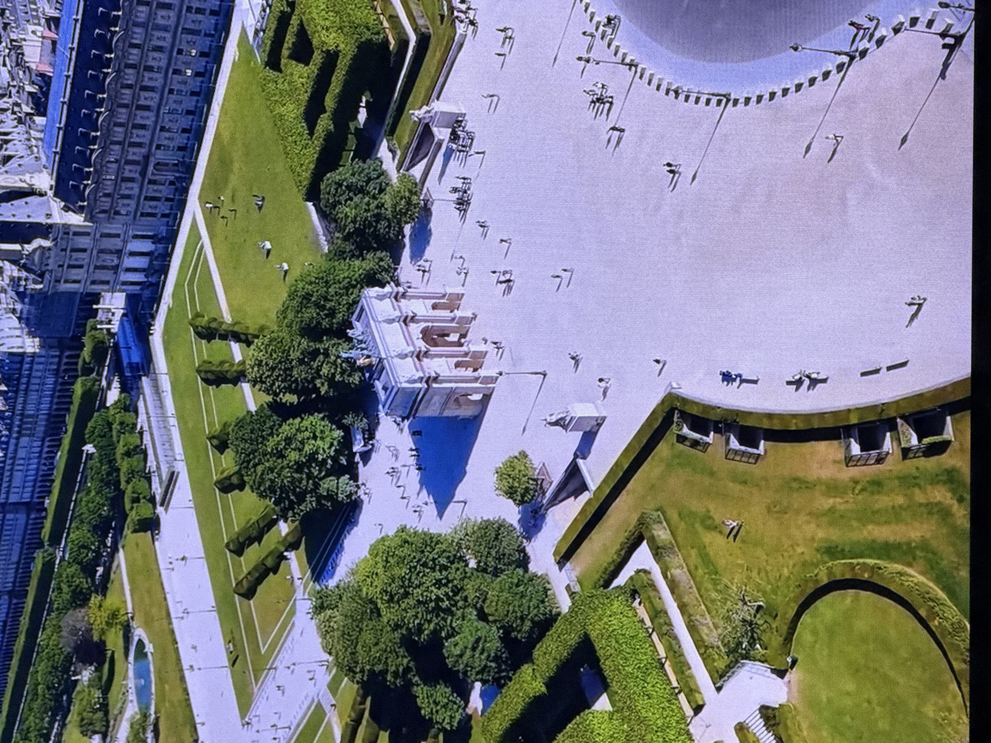 The image is an aerial view of a meticulously landscaped formal garden, likely in Dubai, United Arab Emirates, given the context. The scene depicts a large, open, paved plaza bordering a lush green garden area with manicured hedges and mature trees. Several individuals are scattered across the plaza and within the garden, appearing to be leisurely strolling or taking in the surroundings.
The time of day appears to be mid-morning or early afternoon, with distinct shadows cast by the structures, trees, and people, suggesting bright, sunny weather. The architectural style of the buildings visible in the background, particularly the multi-story structure with numerous windows and balconies, hints at a European influence, possibly a palace or a grand estate. The garden itself features geometric patterns with trimmed hedges forming pathways and borders, a central fountain or pond, and carefully shaped trees.
Notable details include the contrast between the expansive light-colored plaza and the vibrant green of the garden. The people are small figures in the vast landscape, emphasizing the scale of the garden and plaza. There is no visible text in the image. The overall impression is one of tranquility and order, characteristic of a well-maintained public or private garden. The activity is primarily passive enjoyment of the space, such as walking and socializing.