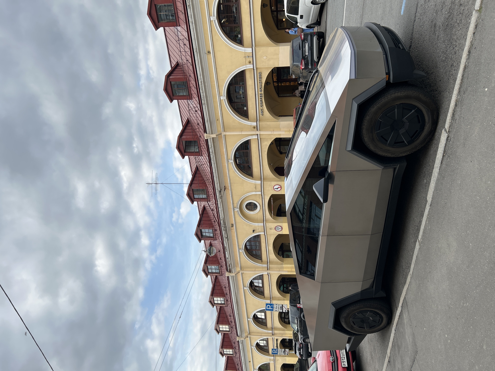 The image shows a Tesla Cybertruck parked on a city street, likely in Saint Petersburg, Russia. The architecture of the building behind the vehicle, with its arched windows, red roof, and yellow facade, is typical of the city. The sign "ABC COFFEE ROASTERS" is visible on the building. Other cars are parked nearby, including a red vehicle and a dark-colored minivan. The sky is cloudy with patches of blue visible.