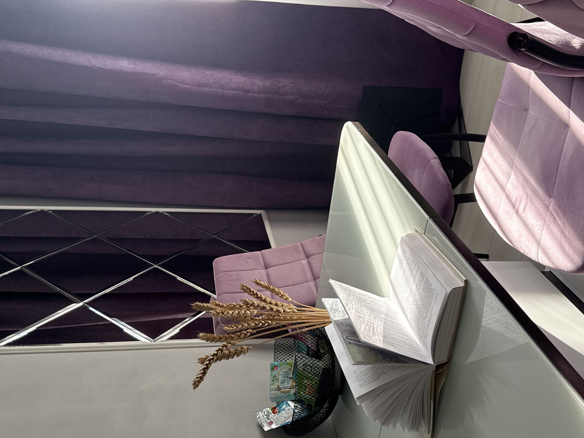 The image depicts an indoor scene featuring a desk with an open book and a vase of wheat stalks. A black mesh basket filled with small packaged items sits on the desk. Two plush purple chairs are visible, one partially obscured by the desk. In the background, a wall is adorned with a diamond-patterned mirror and a deep purple curtain. The scene is illuminated by natural light casting shadows across the desk and chairs. The location is not specified as being in a city.