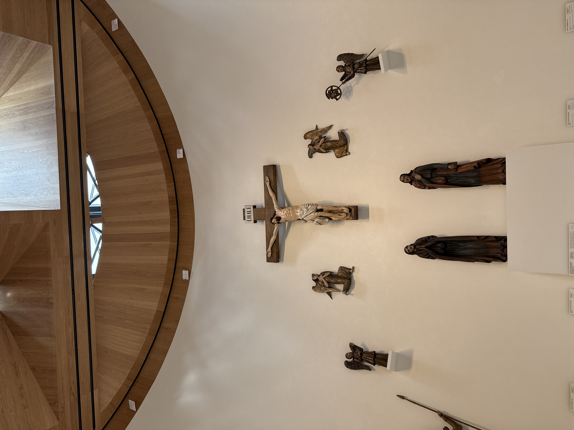The image shows a view of the Perm State Art Gallery in Perm, Russia. A wooden, curved staircase is visible on the left side. On the right is a white wall displaying wooden sculptures and religious art. A central crucifix is featured, surrounded by other figurines. There are placards and labels near some of the sculptures, likely providing descriptions or information.