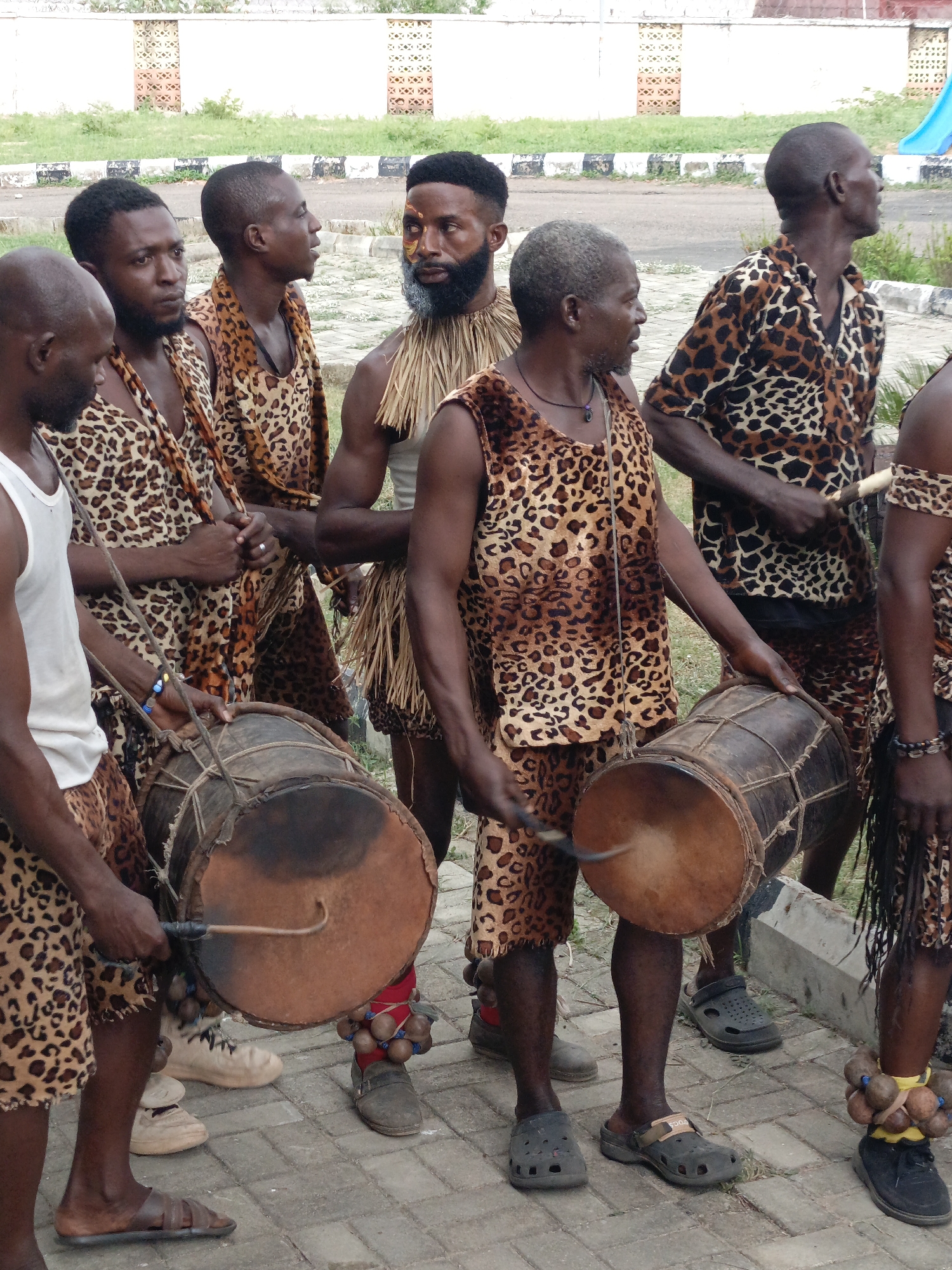 A medium-shot depicts seven men standing together outdoors in Akwanga, Nigeria. The men are positioned on a paved surface with a low concrete structure at the edge. Behind them is a grassy area and a white building with patterned openings. The men are predominantly dark-skinned, with varying ages and hairstyles. Most of the men are wearing leopard-print attire, including vests, shirts, and shorts. One man is wearing a white tank top, and another has a collar made of straw-like material. Several of the men are holding and playing traditional drums. One man has white paint on his face. The men are arranged in a semi-circle, with some looking to the side and others looking forward. The lighting is natural, and the overall impression is of a cultural performance or gathering.