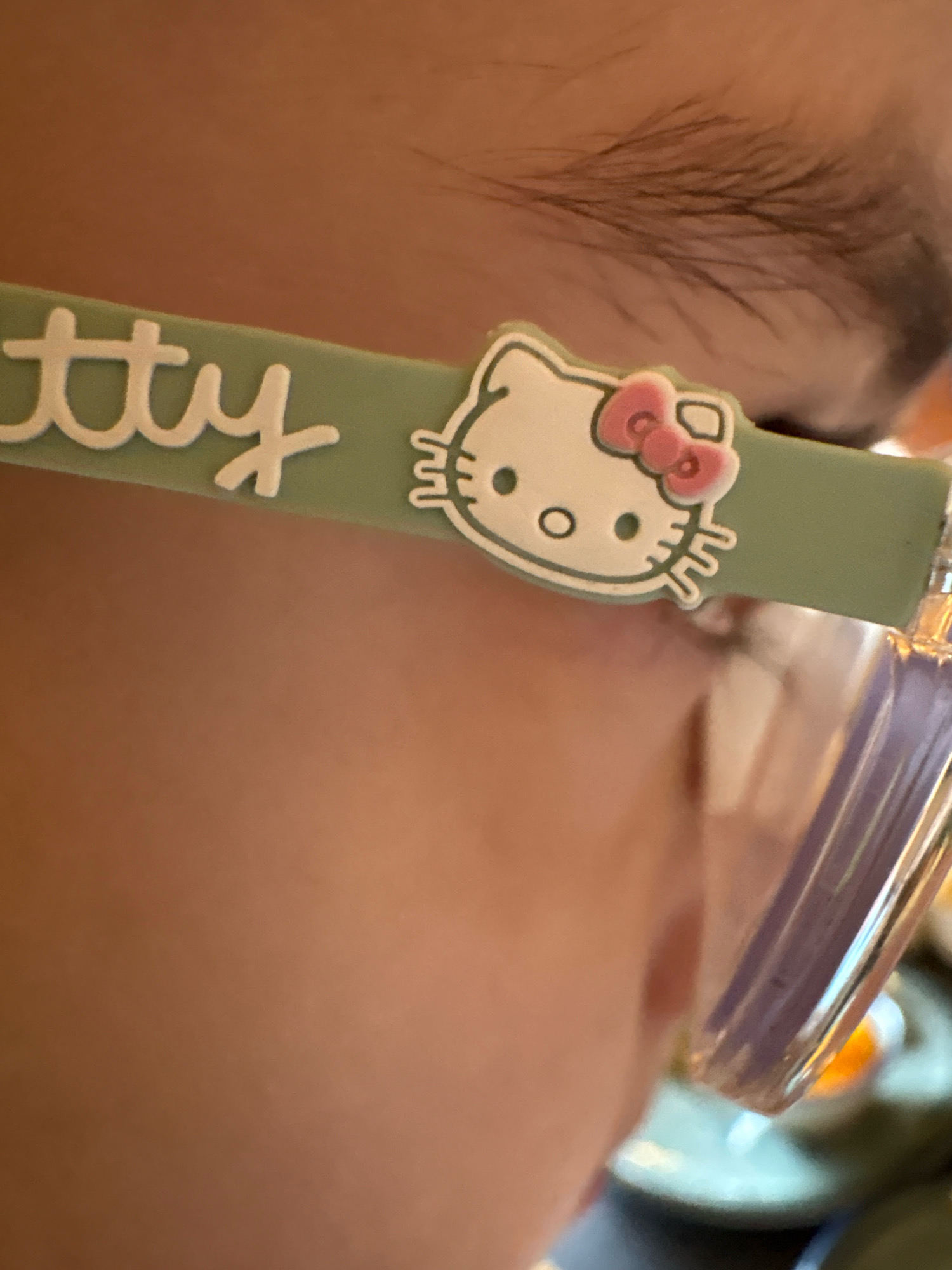 The image shows a close-up of a person's face, partially obscured by a light green object with "tty" written in white cursive letters and a white Hello Kitty charm with a pink bow. The person appears to be wearing glasses, with the top of the glasses frame visible. The background is blurred, but suggests an indoor setting with a hint of orange light. The location is not specified in the image or context.