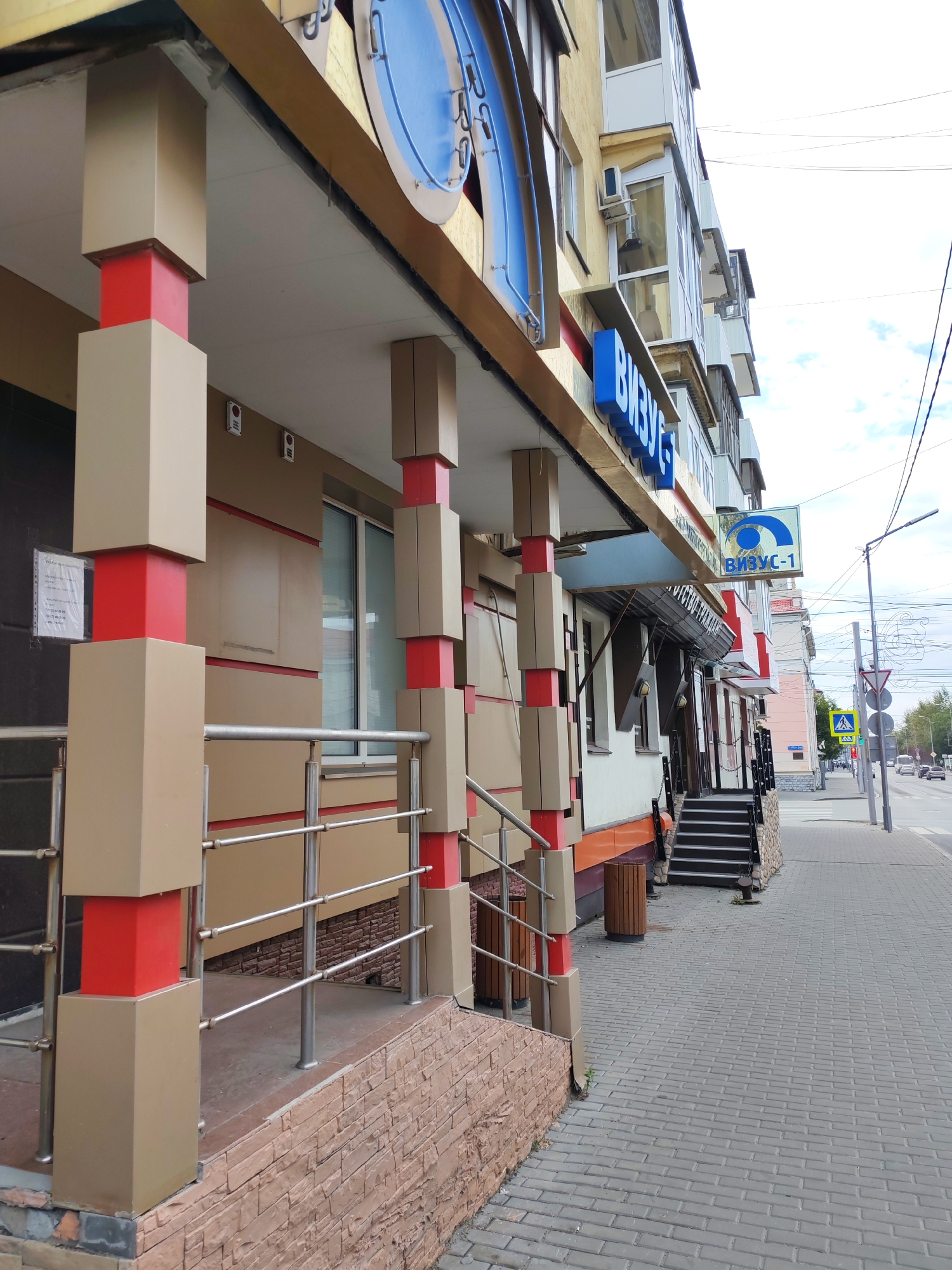 This image shows the exterior of a building in a city. The building has a modern facade with beige and red accents. There is a covered walkway supported by square pillars, some of which are red in the middle section. A metal railing runs along the walkway. Several windows and doors are visible. A blue sign with Cyrillic text is mounted on the building. Further down the street, there is a crosswalk and traffic signals. The sidewalk is paved with grey bricks, and a street with cars is visible in the background. The sky is partly cloudy.