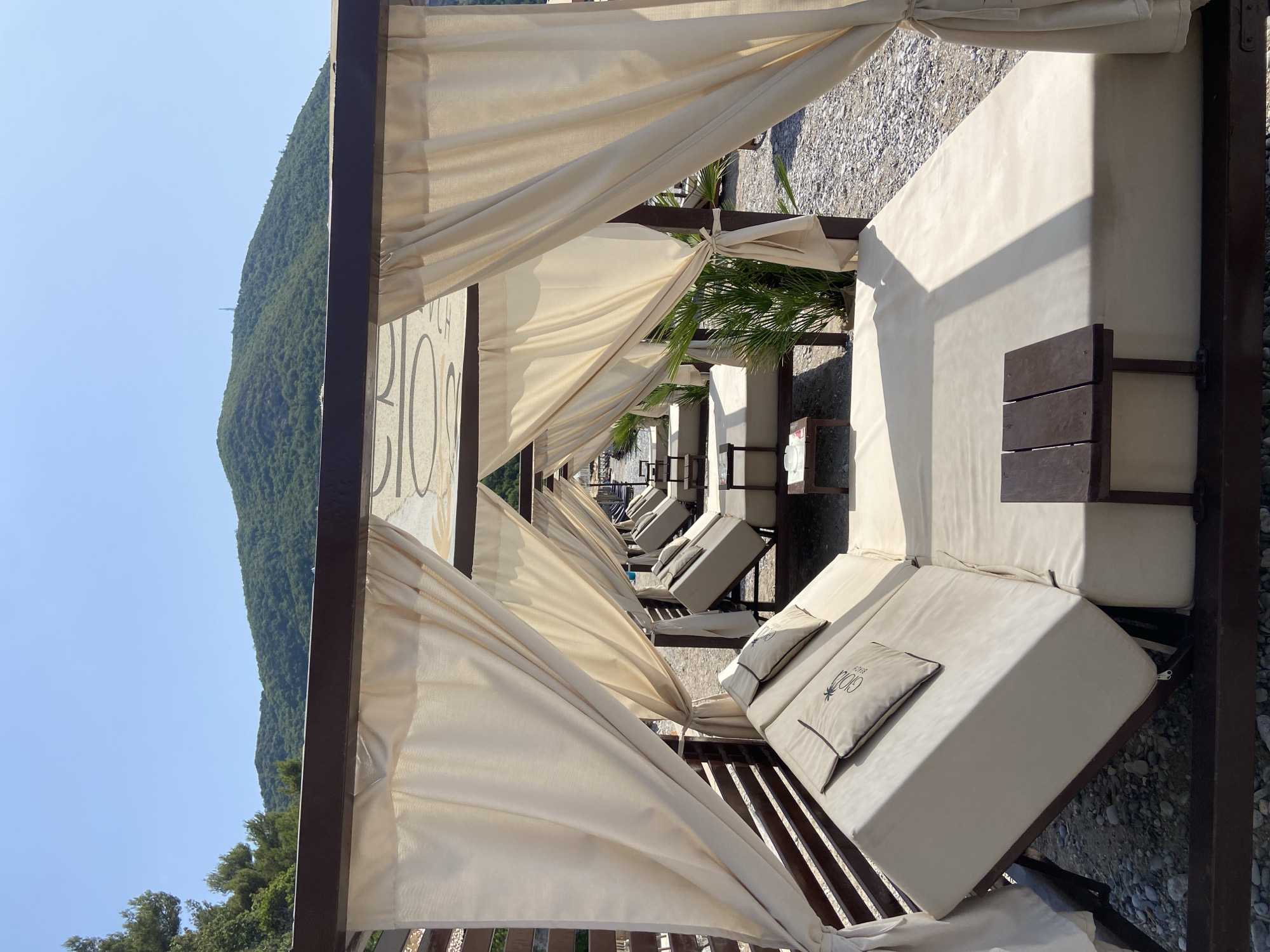 This is a vertical, eye-level, outdoor, medium shot of several private cabanas on a pebbly beach. The cabanas are made of dark wood and have beige curtains that hang down from the open sides. Inside each cabana are large, beige upholstered daybeds with beige pillows. The pillows on the daybeds in the foreground and middle ground have a palm tree logo with the words "GOLD BEACH" below it. Small, dark wood tables are positioned in front of the daybeds. In the background, lush green hills rise under a clear blue sky. The beach appears to be located in Prijevor, Montenegro, as indicated by the "GOLD BEACH" logo, a known establishment in that area. There are no people visible in the image.