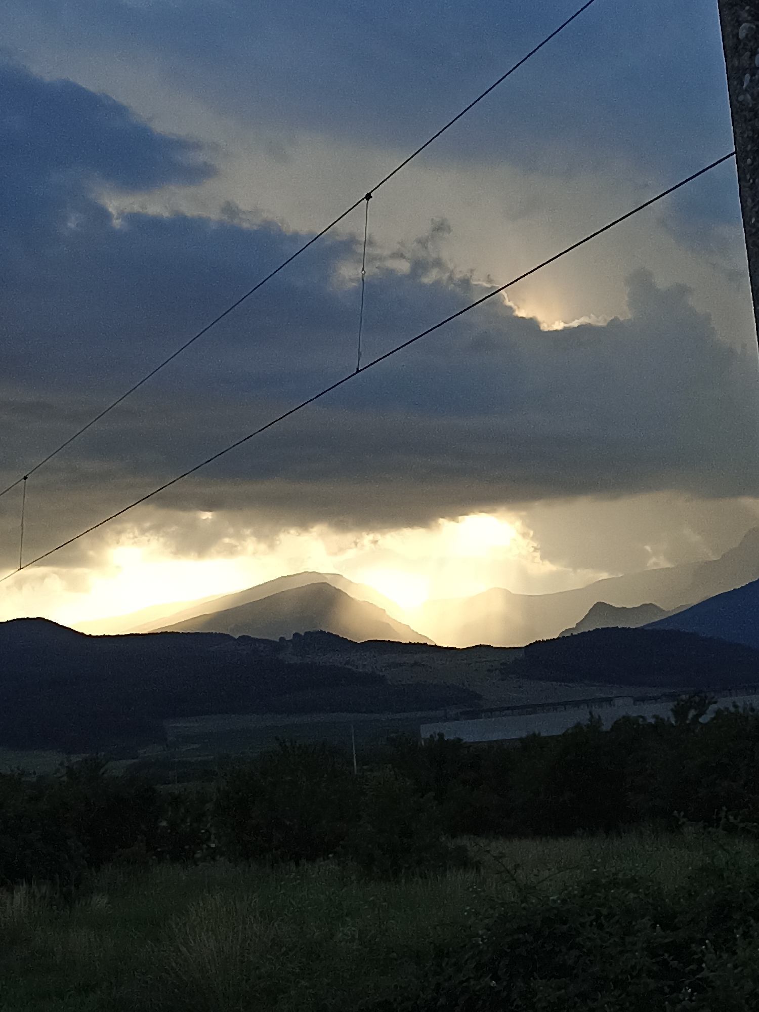 The image shows a dramatic sunset over a mountainous landscape. The sky is filled with dark, heavy clouds, but shafts of sunlight are breaking through, illuminating the peaks of the mountains in a golden light. The foreground is dominated by dark silhouettes of trees and vegetation. A power line stretches across the upper part of the image, casting faint shadows. The scene evokes a sense of natural beauty and perhaps a hint of impending weather.
