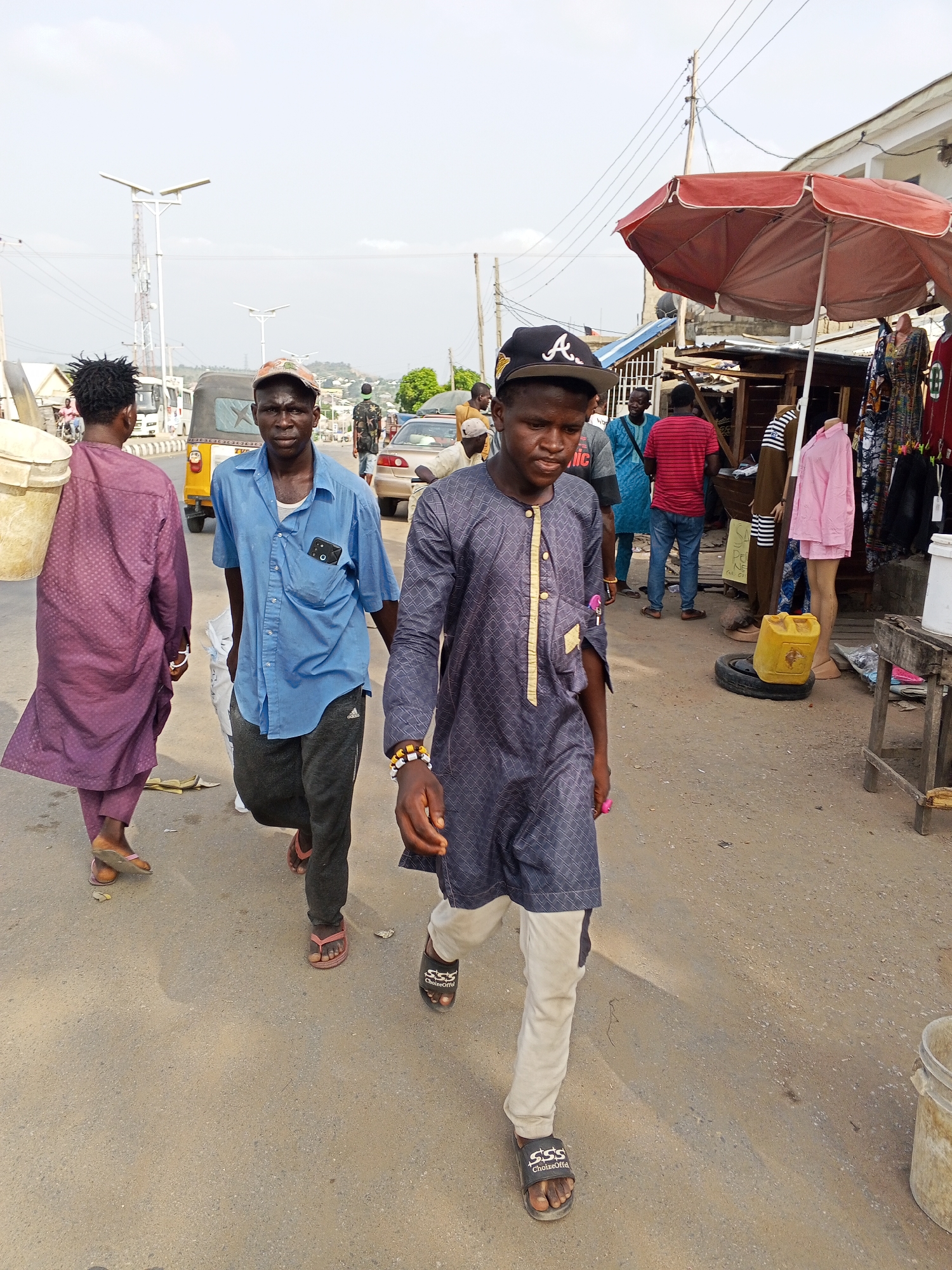 The image shows a street scene in Zango-ndaji, Nigeria. Three men walk along an asphalt road. The man in the foreground wears a patterned grey tunic, light grey trousers, a black baseball cap with the letter "A" on it, and sandals. The man to his left wears a blue shirt, dark sweatpants, and sandals. The third man, furthest left, wears a purple robe and carries a large bucket over his shoulder. Behind them, other people are present, some appearing to be shopping at roadside stalls under a red umbrella. A yellow auto-rickshaw, a parked car, and various buildings are visible in the background. Overhead power lines stretch across the sky.