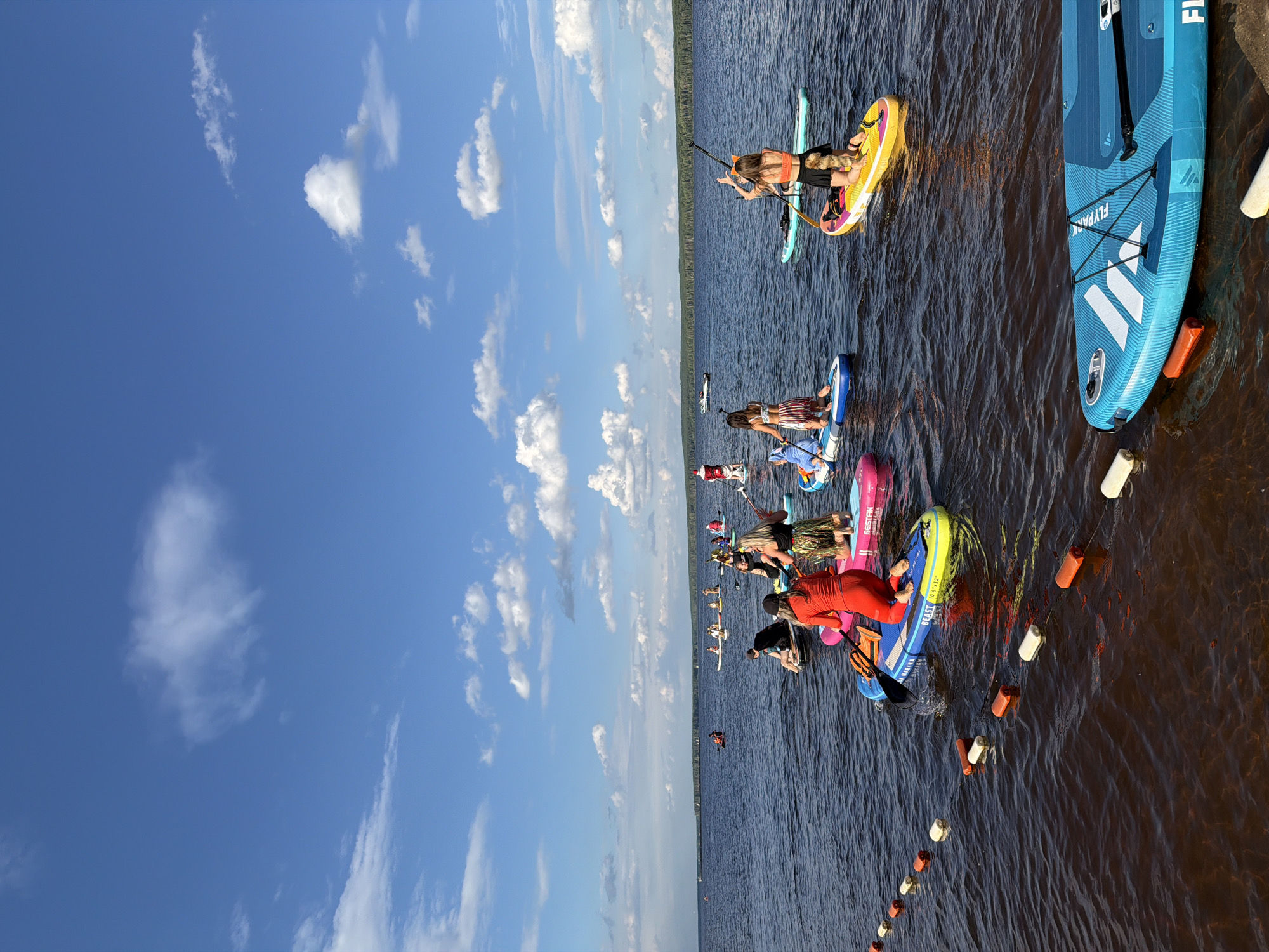 A group of people are paddleboarding on a lake on a sunny day. The sky is blue with scattered white clouds. In the foreground, a bright blue paddleboard is resting on the shore, with a paddle secured to its deck. Orange and white buoys are spaced in a line in the water near the shore. Further out on the lake, several individuals are on paddleboards, some standing and some kneeling. One person is dressed in a red outfit, while another wears a Santa Claus costume. The background shows a treeline and the horizon. The scene appears to be at the Kama River embankment in Chaikovsky, Russia, a location known for its water activities.