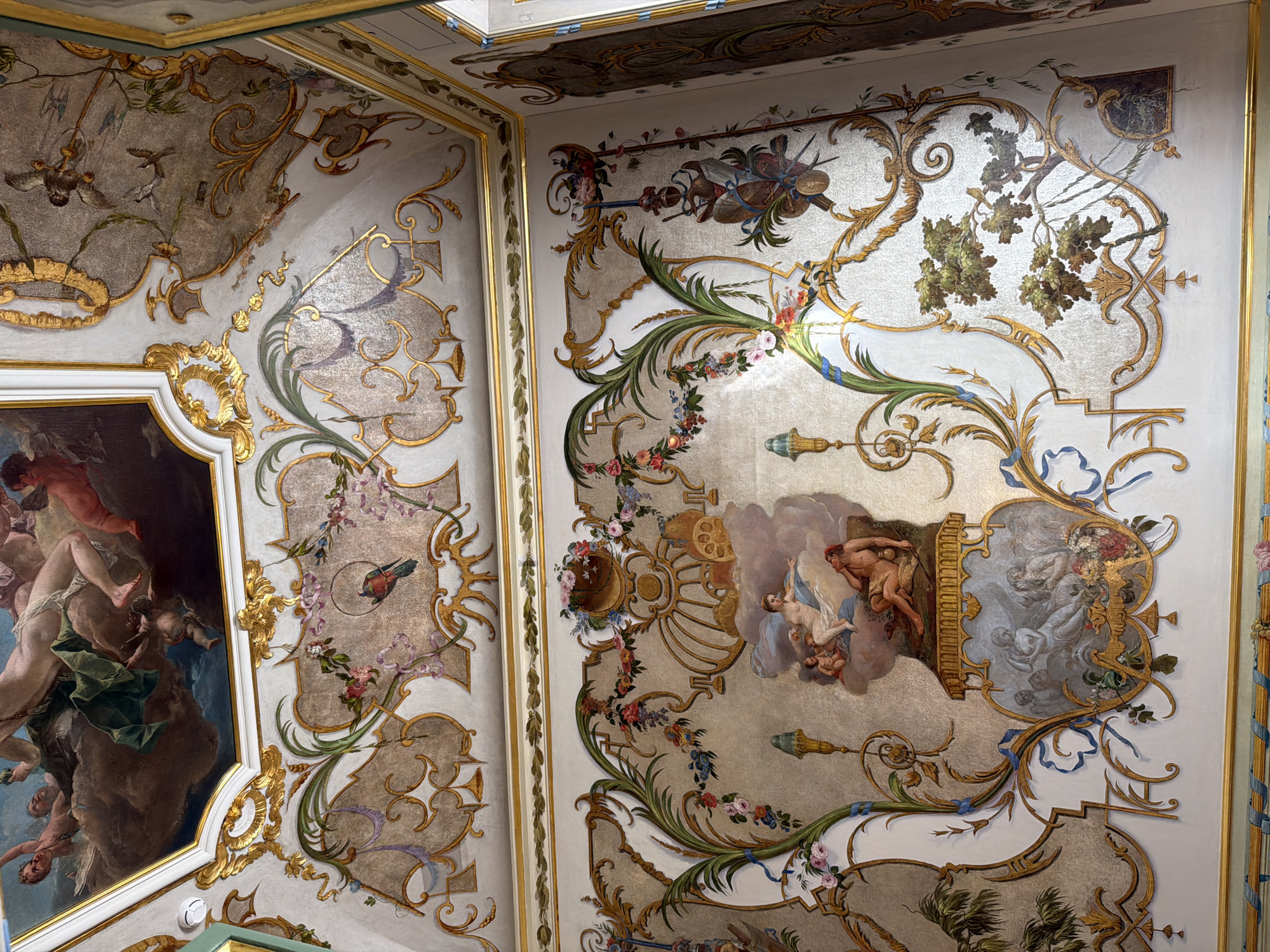 The image shows the interior of a richly decorated room with ornate gilded stucco work and painted murals on the walls and ceiling. The ceiling features a large painting depicting figures in the sky, possibly mythological or allegorical. The walls are adorned with elaborate scrolls, floral motifs, and smaller painted scenes. One prominent mural on the wall depicts several cherubic figures surrounding a reclining male figure and a female figure in the clouds, possibly a scene from classical mythology. Below this, another smaller framed painting features figures in a pastoral setting. The overall aesthetic is opulent and suggests a historic or grand residence. The location is stated as Unknown, Unknown.
