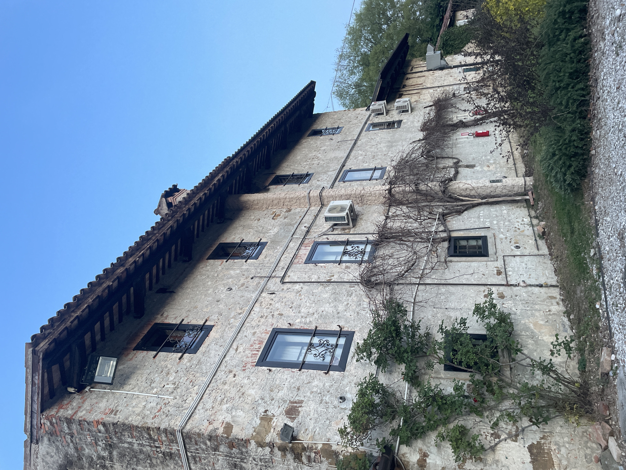 The image depicts the exterior of a multi-story building in San Lorenzo, Italy. The building has a stone facade, windows with decorative metal grilles, and a sloping tiled roof with wooden supports. There are visible air conditioning units mounted on the exterior walls. Plants and vines are growing against the building, obscuring some of the facade. A gravel path is visible on the right side of the building. The sky is clear and blue. The building appears to be old, showing signs of wear and aging.