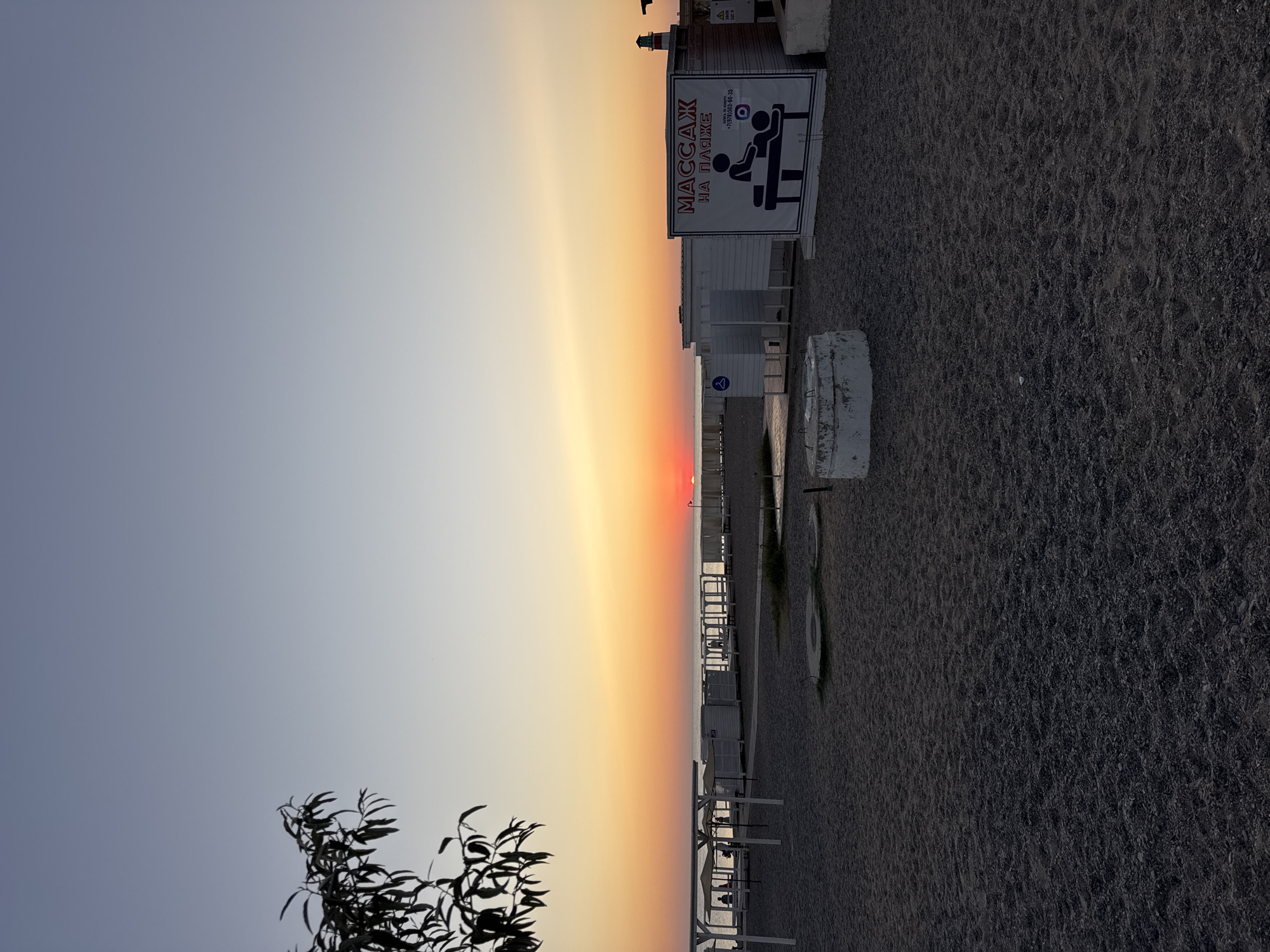 The image depicts a beach scene in Novofedorovka, Ukraine, during sunset. The sky transitions from a light blue at the top to a vibrant orange and red hue near the horizon, where the sun is setting over the sea. The beach is covered in dark gravel, and there are rows of white changing rooms and cabanas. In the foreground, a concrete structure is visible. A sign with the text "MASSAZH NA PLAZHE" (Massage on the Beach) and an illustration of a massage is attached to one of the structures. A tree with dark leaves is silhouetted against the sky in the upper left corner. Several figures, appearing as silhouettes of people, are scattered across the beach, some near the cabanas and others further down the shore. The overall atmosphere is calm and serene, characteristic of a beach at dusk.