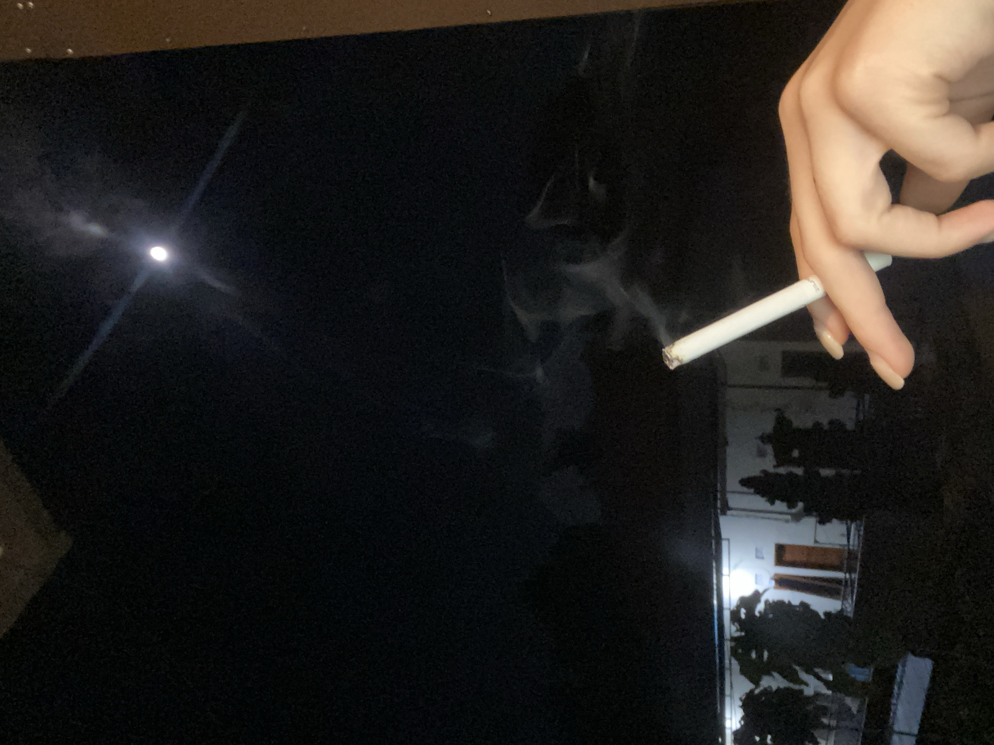 A hand holds a lit cigarette with smoke rising. A dim light source, likely the moon, is visible in the distance on the left. A building with lighted windows is visible in the background. The location, Nusa Dua, Indonesia, cannot be verified from the image.