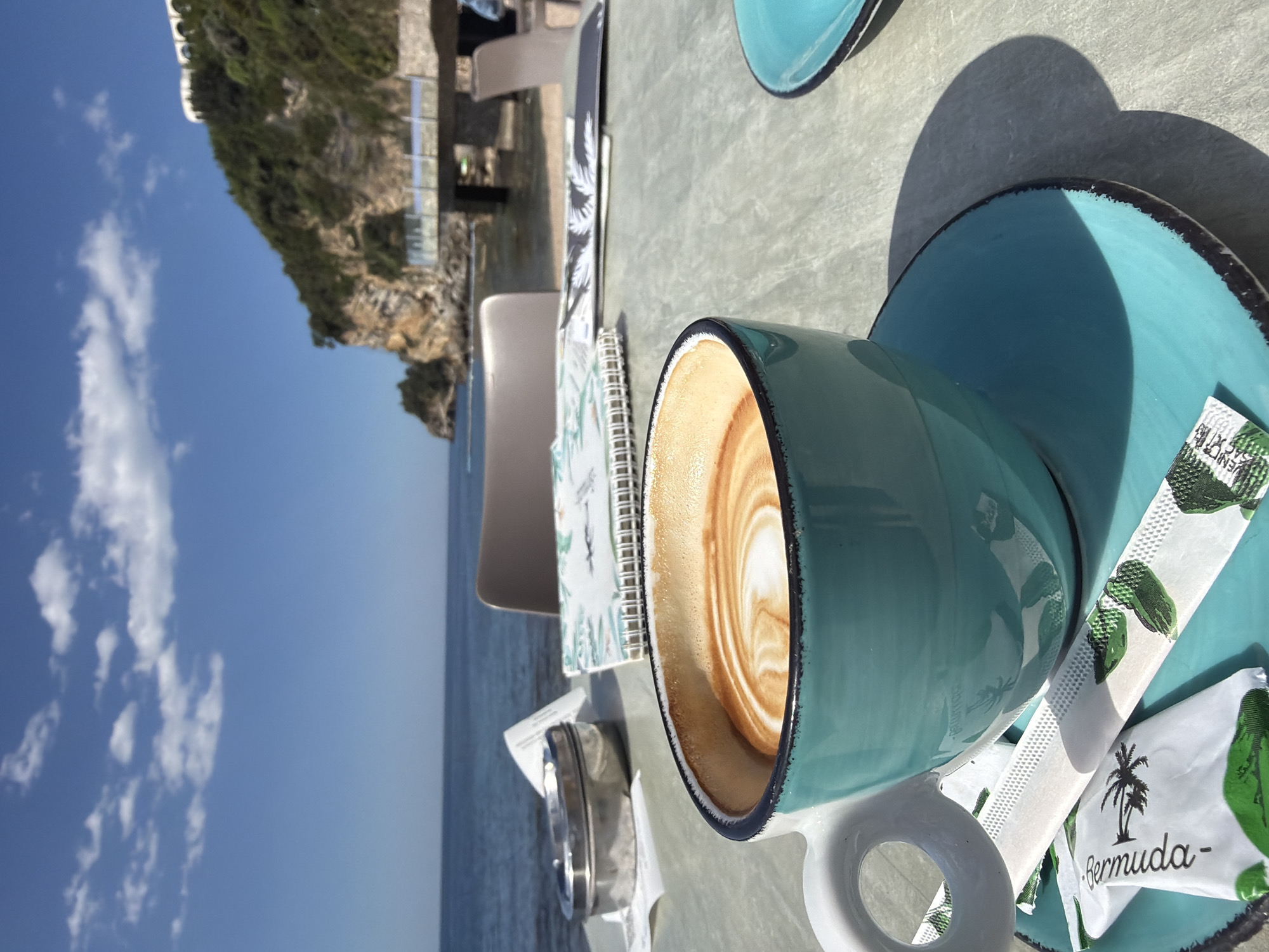 The scene captures a coffee cup on a turquoise saucer, placed on a table at a cafe, with a view of the sea and mountains in Budva, Montenegro. The coffee has latte art. Next to the cup are packets labeled "Bermuda". A notepad and a menu are also on the table. In the background, the clear sky meets the tranquil sea. A mountain range stretches along the coast. Part of a building with a terrace or balcony is visible. The overall environment suggests a serene coastal cafe setting.