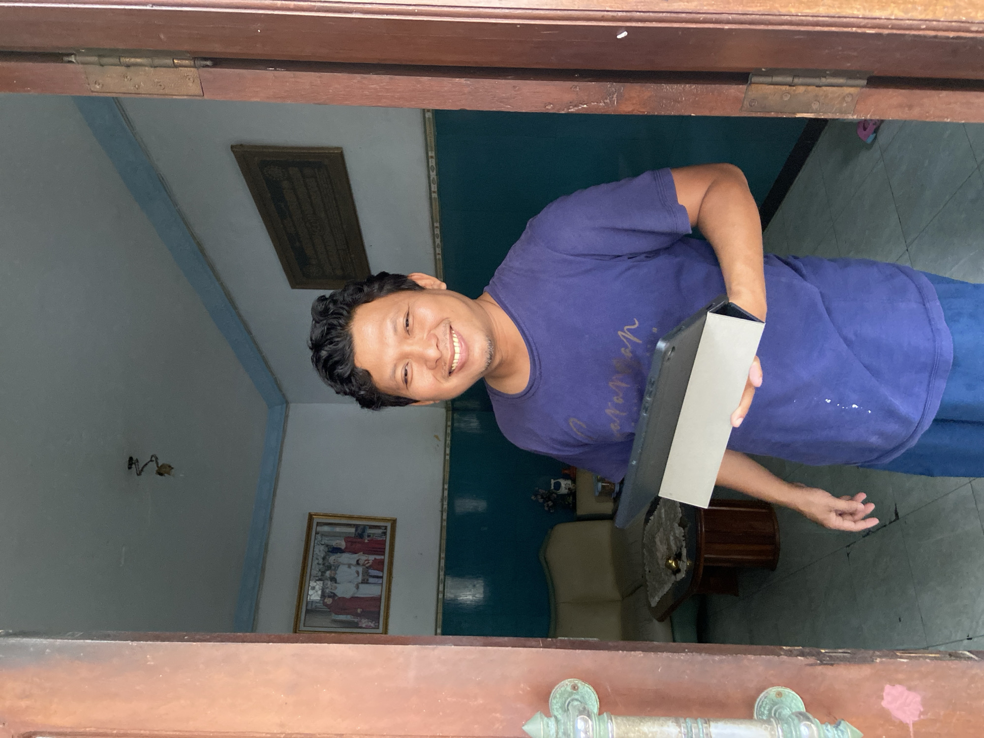 A medium shot through a brown wooden doorway shows an Indonesian man smiling and holding a grey and white box. The man is wearing a purple t-shirt and blue pants. The background reveals a room with white walls, a teal band around the top, and tiled floors. A framed picture hangs on the wall, and furniture, including a light-colored sofa and a small, dark wood table, are partially visible. Based on the presence of Indonesian script on one framed picture, this likely depicts the location in Gelam, Indonesia.