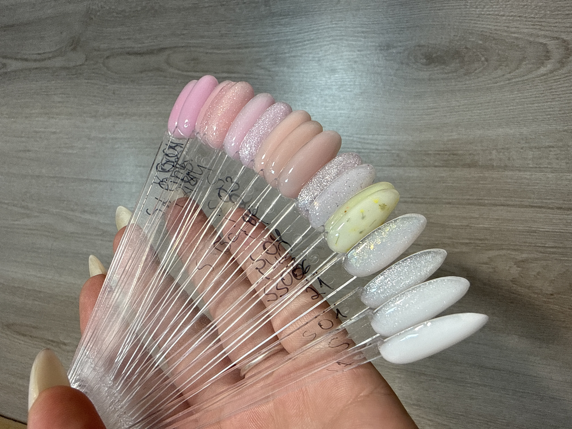 In Burgas, Bulgaria, a hand with light-colored fingernails holds a fan of clear plastic nail polish swatches against a gray wooden surface. The swatches display various shades of pink, white, and a pale yellow with inclusions. Some of the swatches are labeled with black marker, including "404," "804," "901," "21," and "801." The lighting is soft, casting a slight shadow on the wooden background.