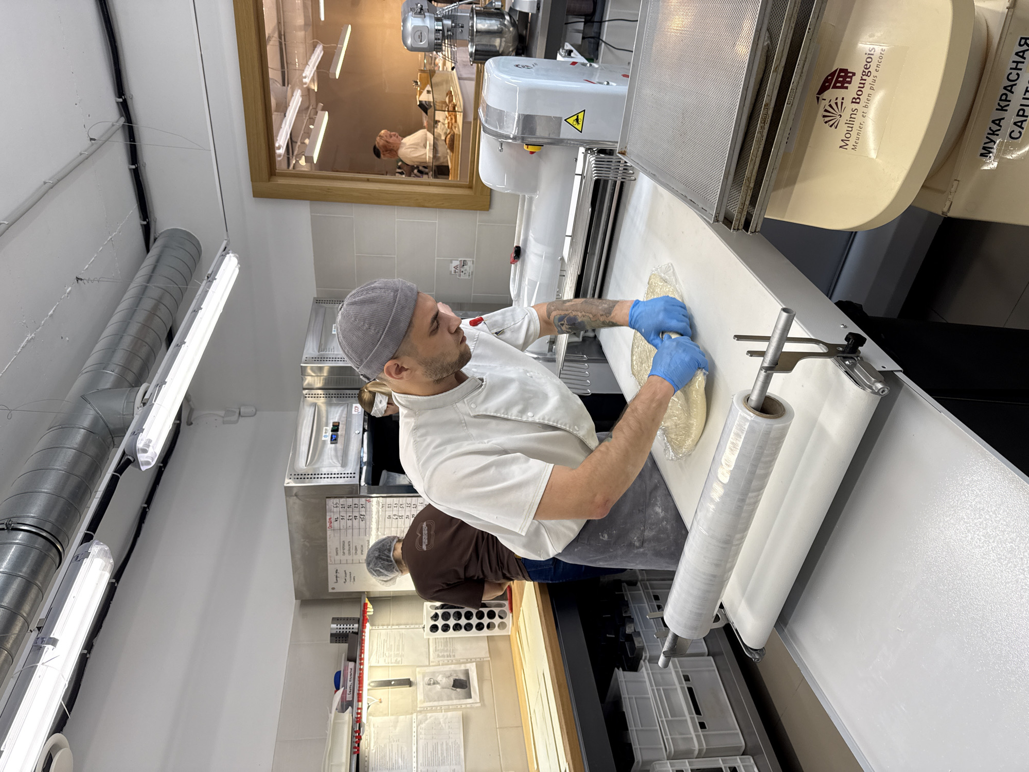 A medium-angle, medium-shot shows a baker preparing dough in a professional kitchen in Vracar Urban Municipality, Serbia. The baker is a light-skinned male wearing a grey cap, a white chef's coat, and a grey apron. He wears blue gloves and manipulates a mass of dough on a white countertop covered in plastic wrap. Behind him, another person, presumably another baker, wearing a hairnet and brown shirt works in the background. Commercial baking equipment is visible, including a dough sheeter and other machinery. The kitchen is well-lit with fluorescent lighting and features stainless steel surfaces. The scene appears to be a clean and orderly food preparation area. Signage on the equipment includes French and Cyrillic text.