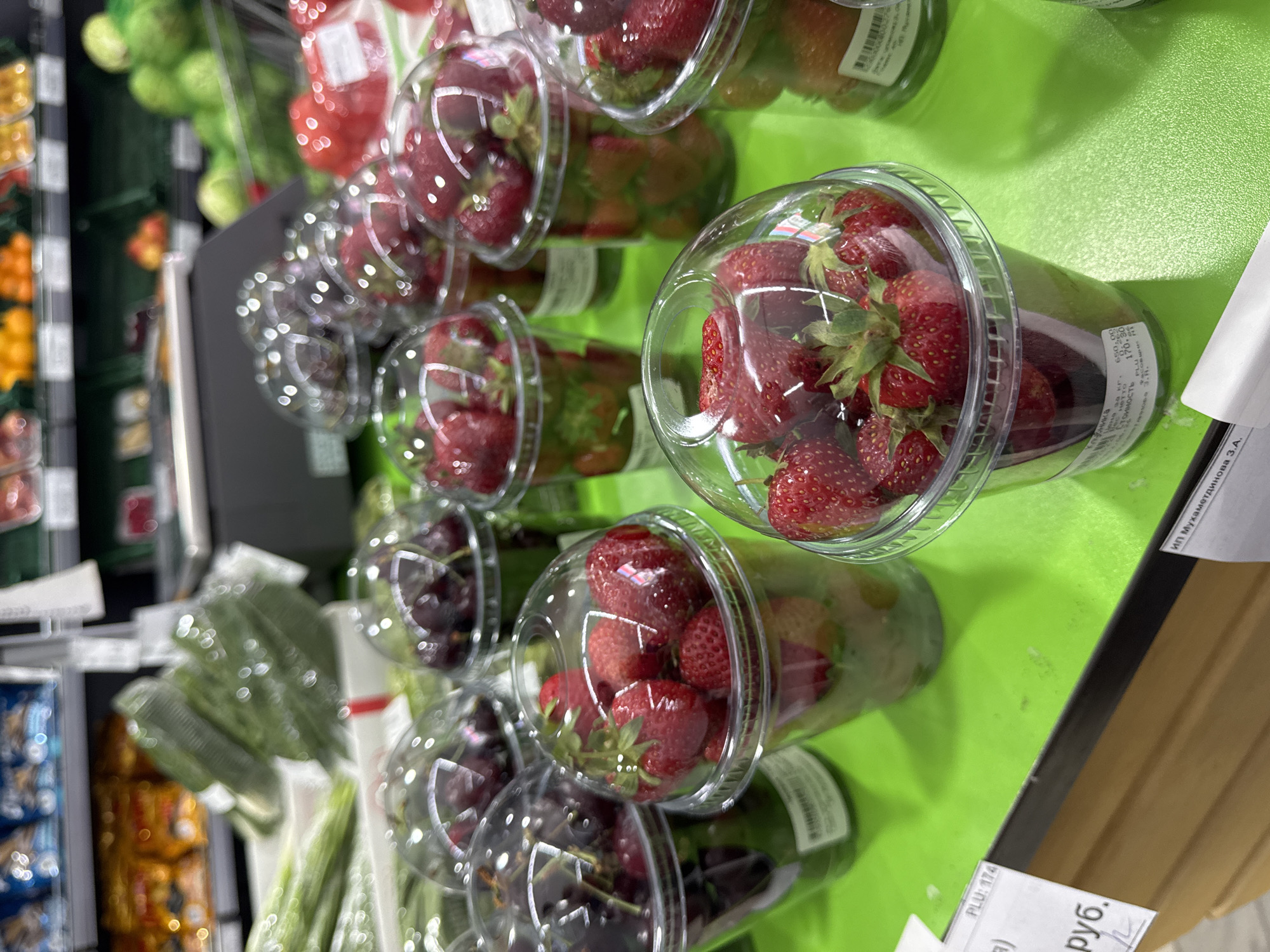 The image shows a close-up of plastic cups filled with fresh strawberries and cherries, displayed on a bright green surface. The products appear to be for sale in a retail environment. In the background, shelves stocked with various produce and packaged goods are visible. Several price tags are attached to the cups and the display surface. One price tag shows "PLU: 174" and another is partially visible with text including "ИТ, Мухаметдинов З.А.". The scene is indoors, likely in a grocery store or market. The lighting is bright, illuminating the vibrant colors of the fruits. There are no people visible in the frame.
