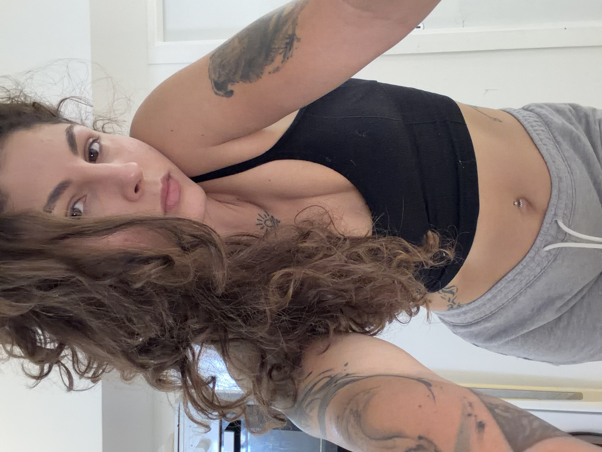 A young woman is captured in a close-up, rotated selfie, appearing to be lying down indoors. She has long, voluminous dark brown curly hair that partially covers her chest and flows into the foreground. Her dark eyes are open, and she looks directly at the camera with a neutral expression. She is wearing a black sports bra or crop top and light grey sweatpants with a white drawstring.
Notable details include multiple tattoos: intricate designs are visible on both forearms, a small sun-like symbol is tattooed on her chest/sternum area, and another subtle tattoo (possibly an arrow) is on her upper abdomen. A small, shiny silver-toned belly button piercing is also visible. The background is a plain, light-colored wall, suggesting an indoor setting with minimal distractions. The lighting is soft and even, indicating an absence of harsh sunlight. No visible text is present in the image. The overall mood is relaxed and casual. This scene is set in Kiryat Bialik, Israel.