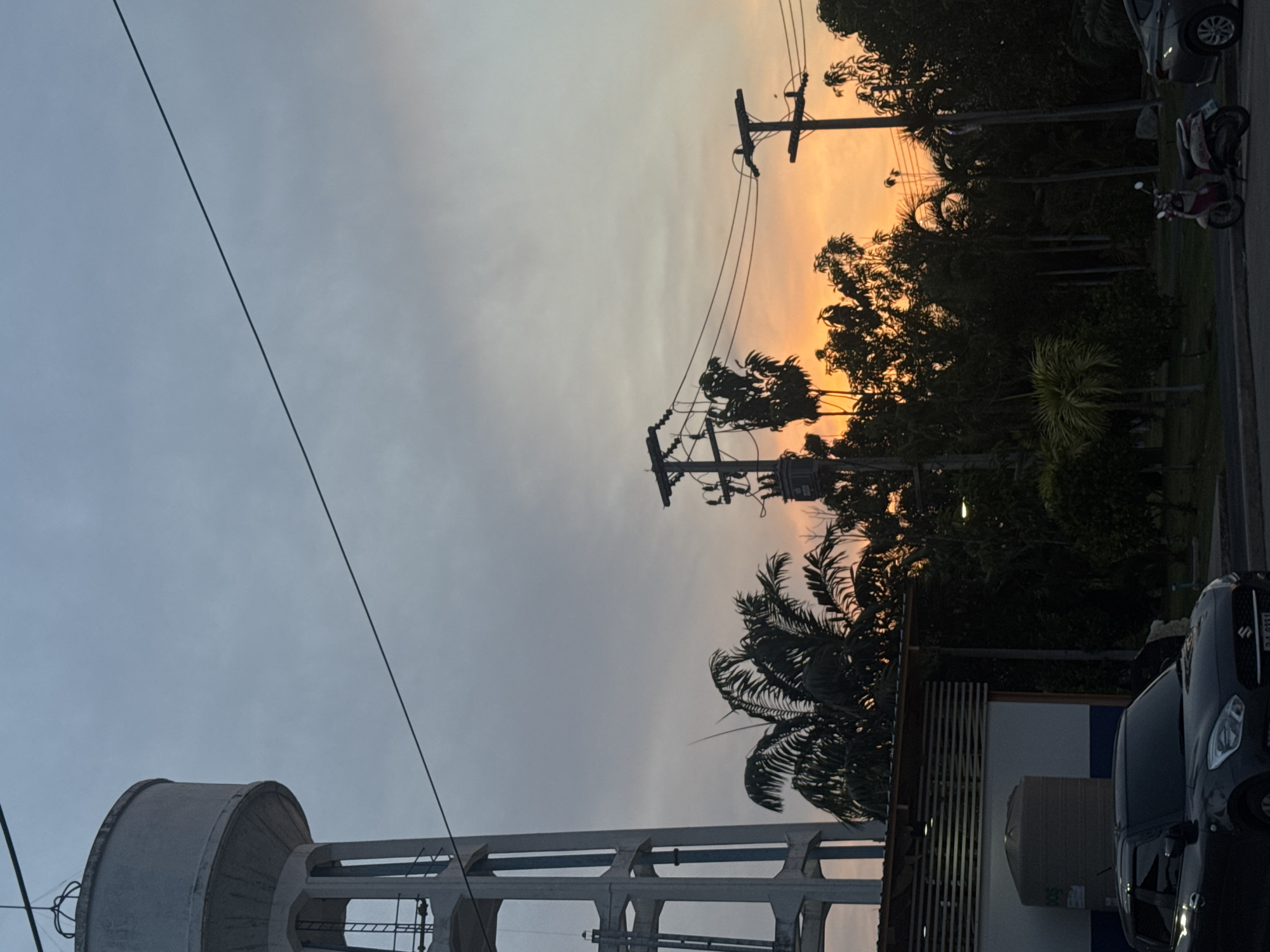 This media file captures an outdoor twilight scene in Pattaya City, Thailand, likely at sunset or sunrise, with a striking sky gradient. The sky transitions from a soft grey-blue at the top to a vibrant orange and yellow on the horizon, backlighting the landscape elements.
Dominating the left side of the frame is a substantial concrete water tower, featuring a large cylindrical tank elevated by a framework of concrete pillars and support beams. Multiple utility poles, laden with a complex web of overhead power lines and cables, stretch across the middle ground, appearing in stark silhouette against the bright sky.
On the right side of the image, a dense canopy of tropical trees, including numerous palm trees, forms a dark silhouette against the luminous horizon, suggesting a rich natural environment. Adjacent to this foliage, elements of a roadside scene are visible: a dark-colored car, possibly a Suzuki, is partially in view, with its license plate "DG 9331" discernable. Further back, a scooter or motorcycle is also parked on the shoulder. Part of a building or structure with a white wall and vertical wooden slats can be seen at the bottom right.
No individuals are present in the photograph. The image conveys a peaceful atmosphere, highlighting the contrast between man-made infrastructure and natural beauty during the golden hour.