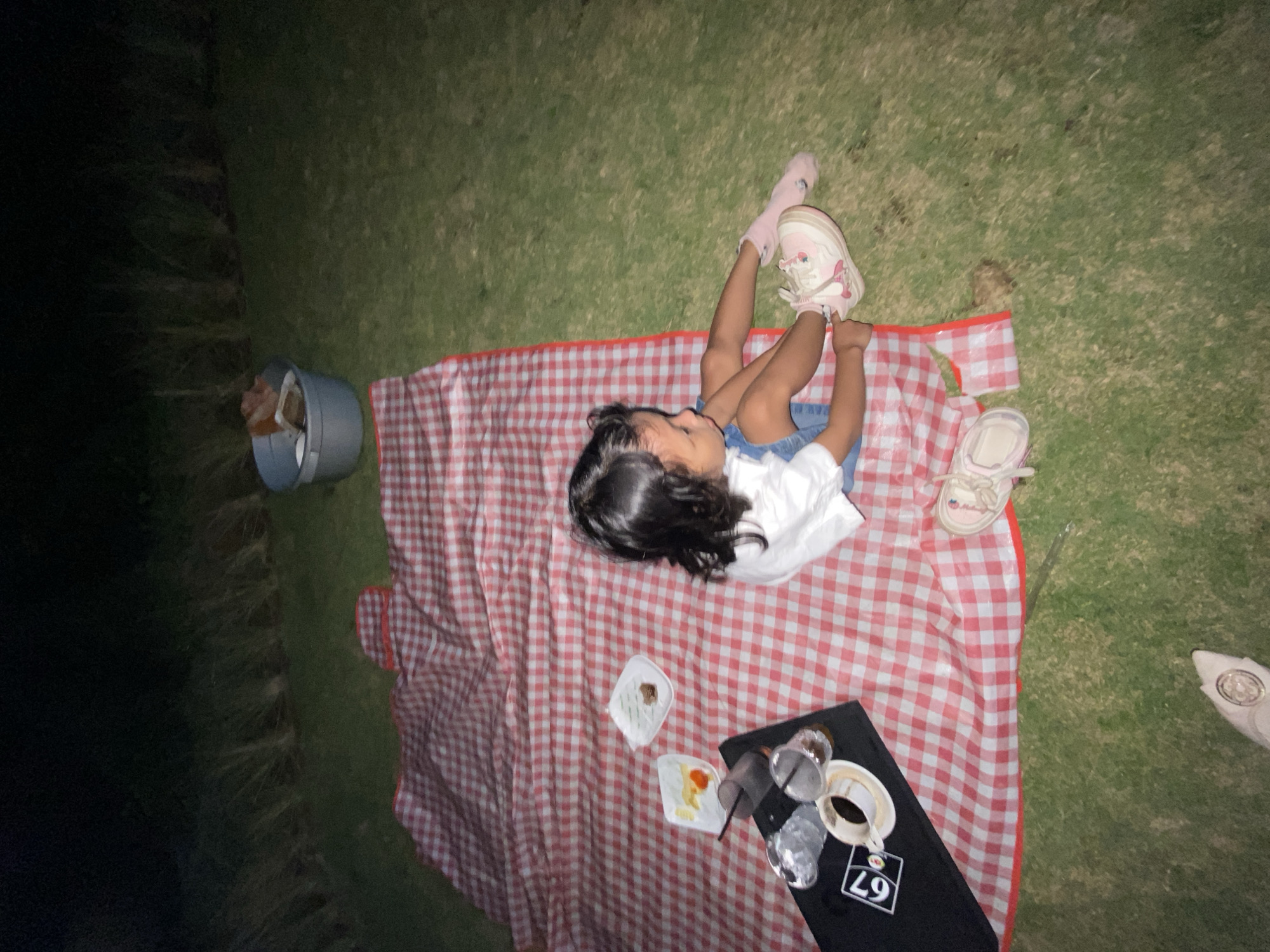 A young girl is lying on a red and white checkered picnic blanket spread on grass. She is wearing a white top, blue shorts, pink socks, and pink and white sneakers. Her dark hair is spread around her head. Two plates with food are on the blanket near her. A black tray with a cup of coffee and some cups is also on the blanket. A gray bucket with some trash is to the left of the blanket. The scene appears to be outdoors, possibly in a park or backyard, and it is nighttime or dusk, as indicated by the low lighting and shadows. The girl's posture and the picnic setup suggest a casual outdoor gathering or a break during an activity. There is some text visible on a white tag on the tray: "L9". The setting is Kesiman, Indonesia.
