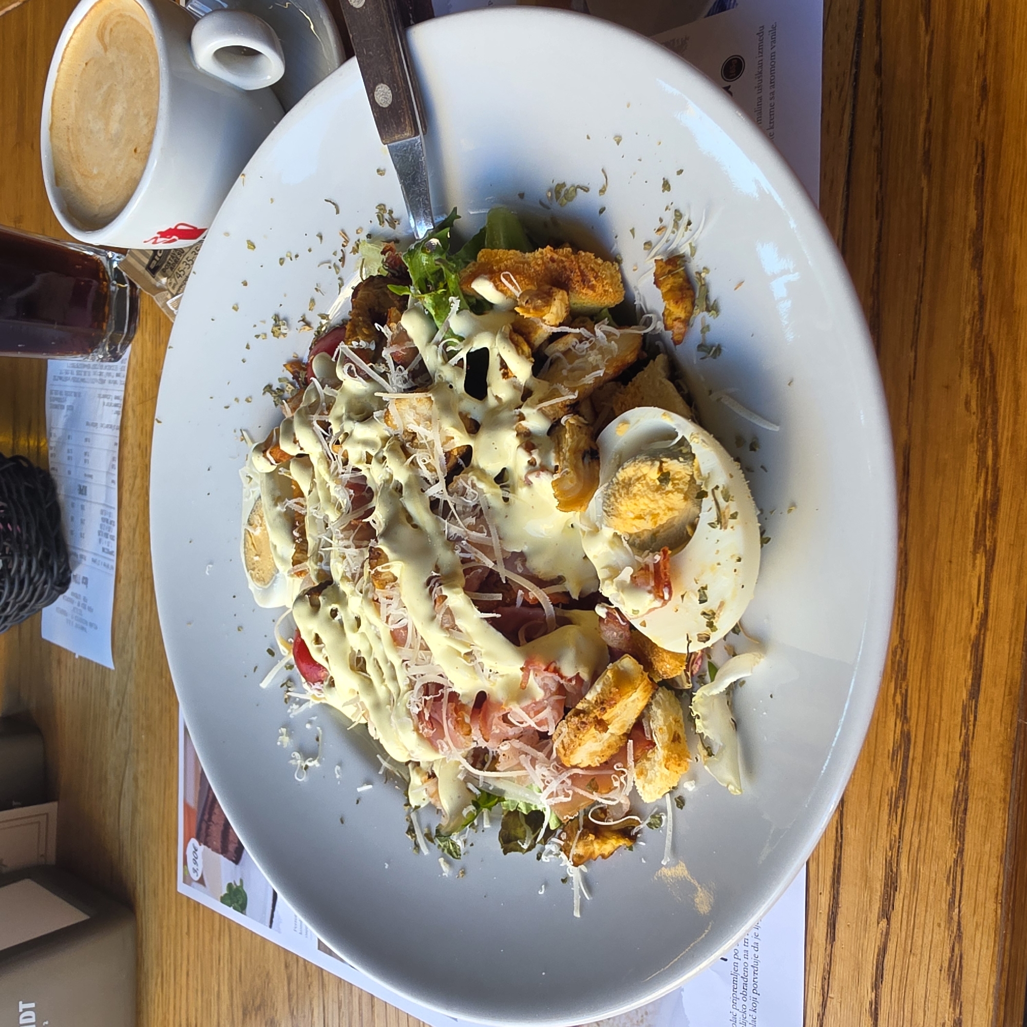 The image shows a close-up of a plate of salad on a wooden table. The salad appears to be a Caesar salad with grilled chicken, croutons, hard-boiled eggs, and shaved parmesan cheese, drizzled with a creamy dressing. There are also some sprigs of fresh herbs sprinkled over the salad. To the right of the salad, a white cup of coffee with latte art is visible, along with a small glass of dark liquid, likely soda or iced tea. In the background, there is a receipt and a black woven basket. The lighting suggests an indoor setting, possibly a restaurant or cafe. The text on the menu visible in the lower left corner is in a Slavic language, and one of the menu items translates to "prepared in a special way." The overall scene is one of a meal in progress. This image is likely from Podgorica, Montenegro, a city known for its blend of traditional and modern cafes and restaurants.