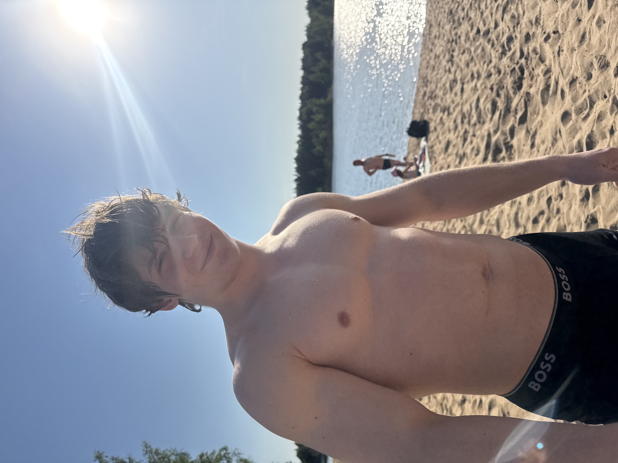 A young man is photographed from a low angle on a beach. He is shirtless, wearing black "BOSS" underwear. The beach has a sandy surface, and there is a body of water behind him with trees lining the opposite shore. Other individuals are visible in the background, some near the water. The sky is bright blue with the sun visible, causing lens flare. The location is identified as a beach in the city of Novyi, Russia.