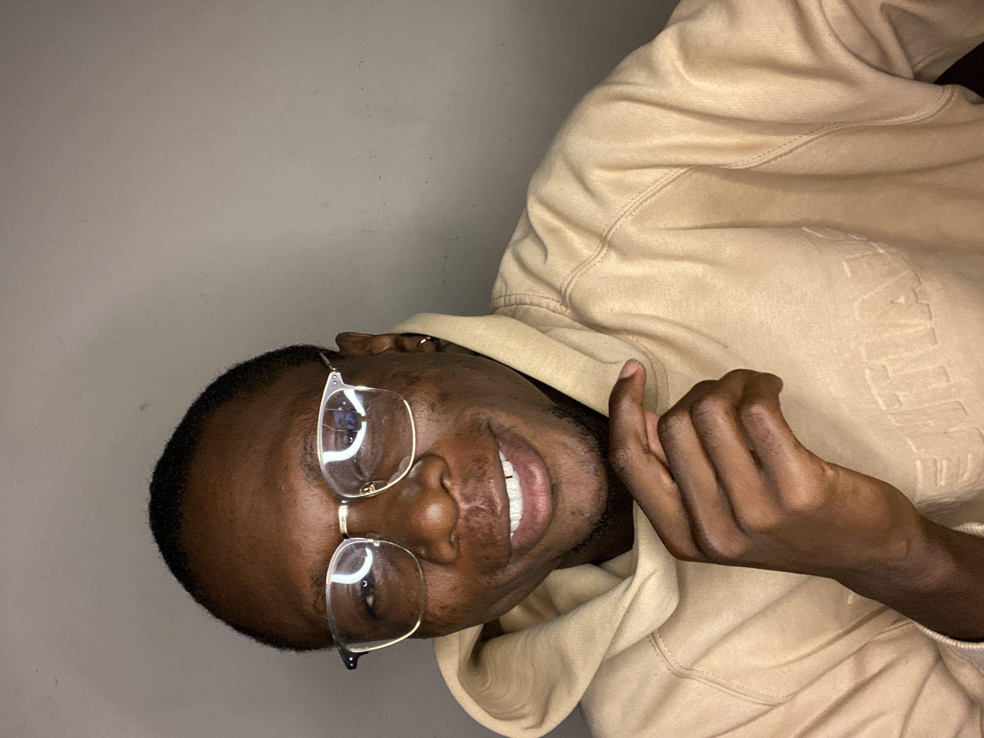 A young Black man is captured in a close-up, chest-up shot, smiling broadly and looking directly at the camera. He wears clear-framed glasses and a light beige or tan hooded sweatshirt. His short hair is neatly cut, and a small earring is visible in his right ear. His left hand, partially clenched, rests against his hoodie on the right side of the frame. The hoodie features faint, embossed text across the chest, which clearly spells out "ESSENTIALS." The background is a plain, light gray wall, indicating an indoor setting with soft, even lighting. The man's bright, open smile conveys a friendly and positive emotion. The photograph is set in Bukuru, Nigeria.