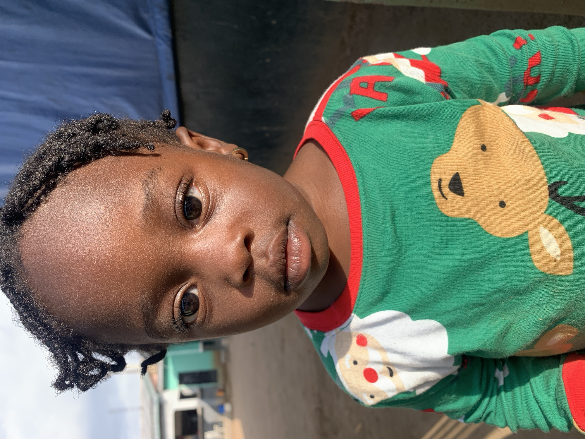 The image, presented on its side with a vertical orientation, features a close-up portrait of a young child, likely female, with dark skin and short, dark, textured hair styled in twists or starter locs. The child's face is central, looking directly at the viewer with wide, expressive eyes and slightly pouted lips, conveying a neutral to serious expression. A small, gold-colored hoop earring is visible on the child's right ear.
She is wearing a vibrant green long-sleeved top with red ribbed trim along the collar and cuffs. The shirt features festive holiday-themed graphics, including a large brown reindeer head with a black nose and eyes, and a cheerful Santa Claus face with a white beard and red hat. Partially visible red text, including a clear 'A', is printed on the green fabric.
The outdoor setting is brightly lit by direct sunlight, casting distinct highlights on the child's skin and suggesting a clear, sunny day, likely mid-morning or early afternoon. The background is softly blurred, revealing a dark blue fabric or tarp-like structure in the upper left and indistinct light-colored buildings or structures with some ground in the lower right. The overall scene captures a candid moment in what is identified as New Stadium, Nigeria.