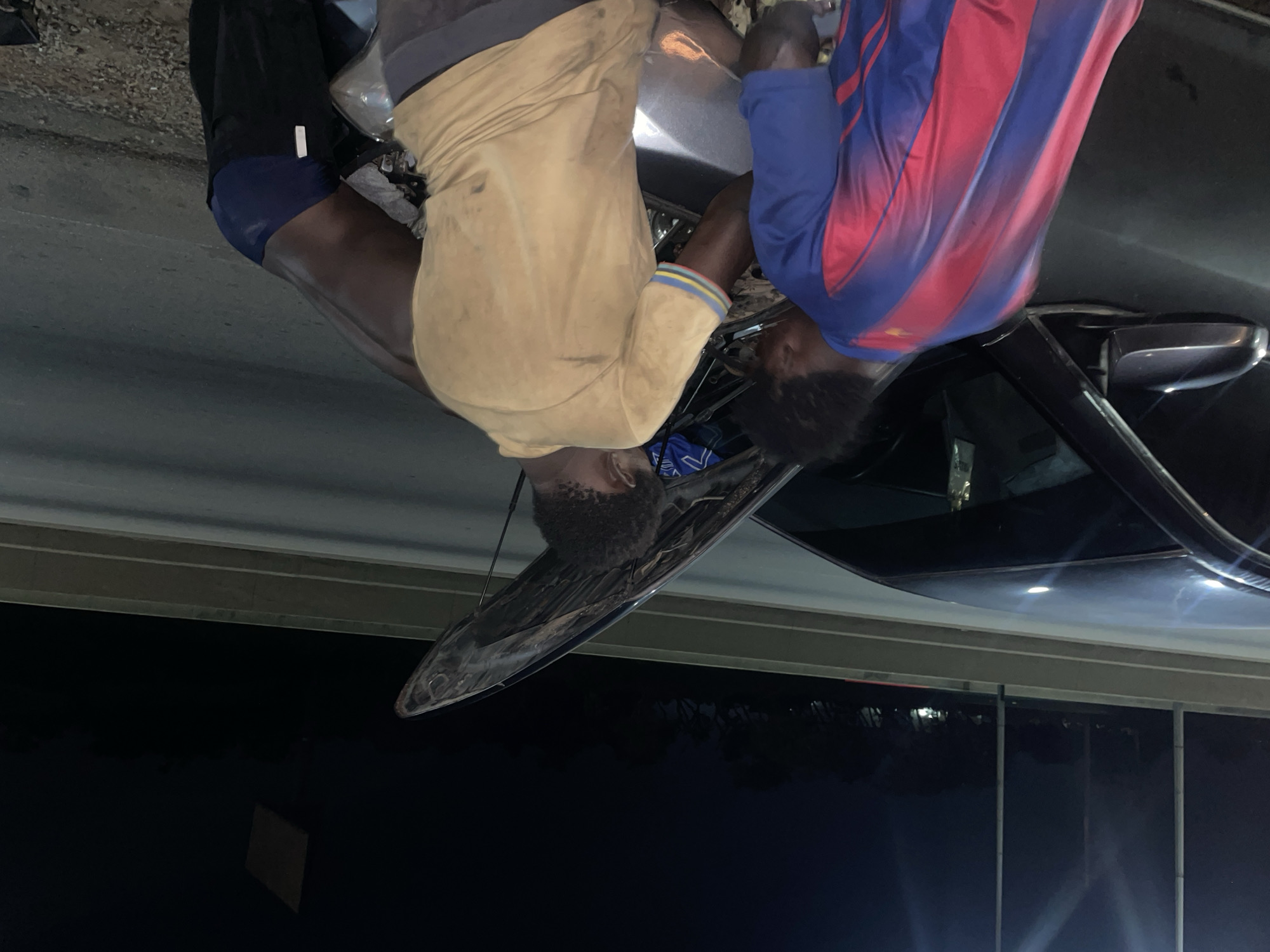 The image shows a group of men working on a car outdoors at night. Two men are visible in the foreground, one wearing a beige shirt and dark pants, and the other wearing a blue and red striped shirt. They appear to be leaning over the hood of a black car, possibly making repairs or modifications. The scene is dimly lit, suggesting it is nighttime, and there are some artificial lights reflecting off the car's surface. The ground appears to be paved, and there's a hint of a building or structure in the background. The men's focus is on the car, and their posture suggests an engaged and perhaps collaborative effort. One man's face is partially visible, showing a determined expression. The overall atmosphere is one of practical work being done in an informal setting. There is no visible text in the image.