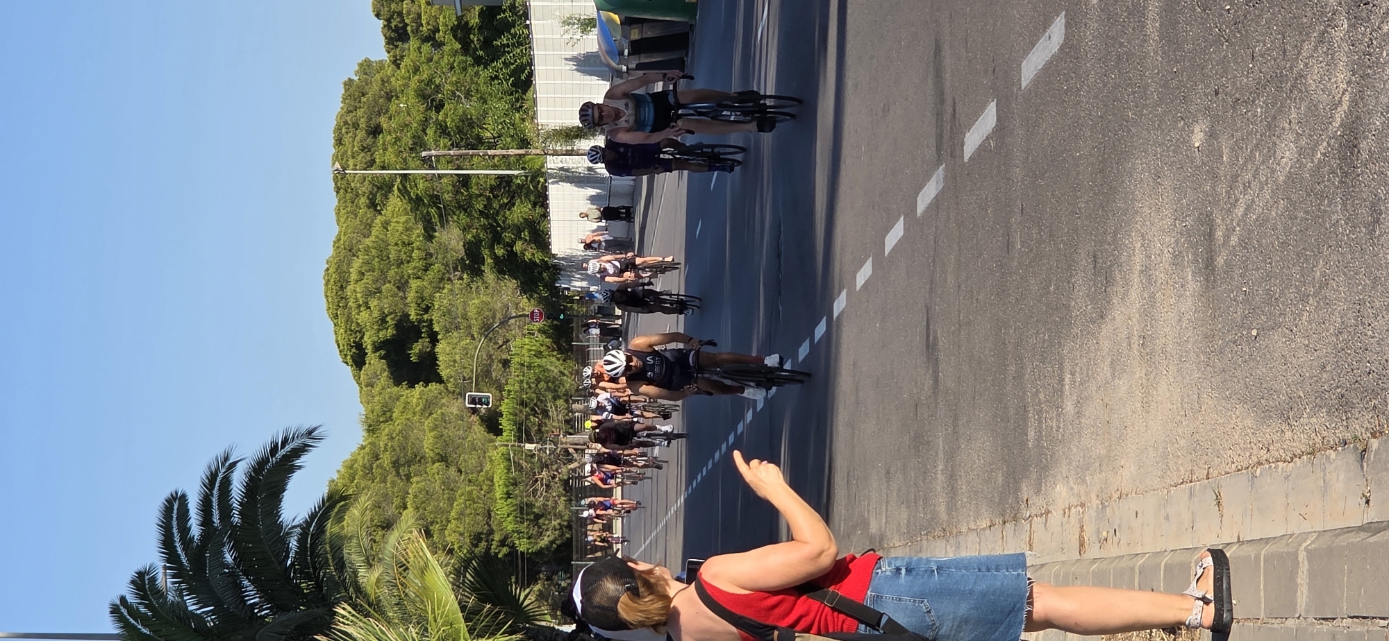In the city of Grao, Spain, a group of cyclists is participating in a race on a sunny day. The cyclists, wearing helmets and racing attire, are riding on a paved road with white dashed lines. A woman in a red top and denim skirt stands on the sidewalk, pointing her finger towards the cyclists. Palm trees and dense green trees line the background, with a clear blue sky above. Traffic lights and a stop sign are visible on the roadside, indicating an intersection ahead. The scene appears to be a typical outdoor sporting event in a coastal area.
