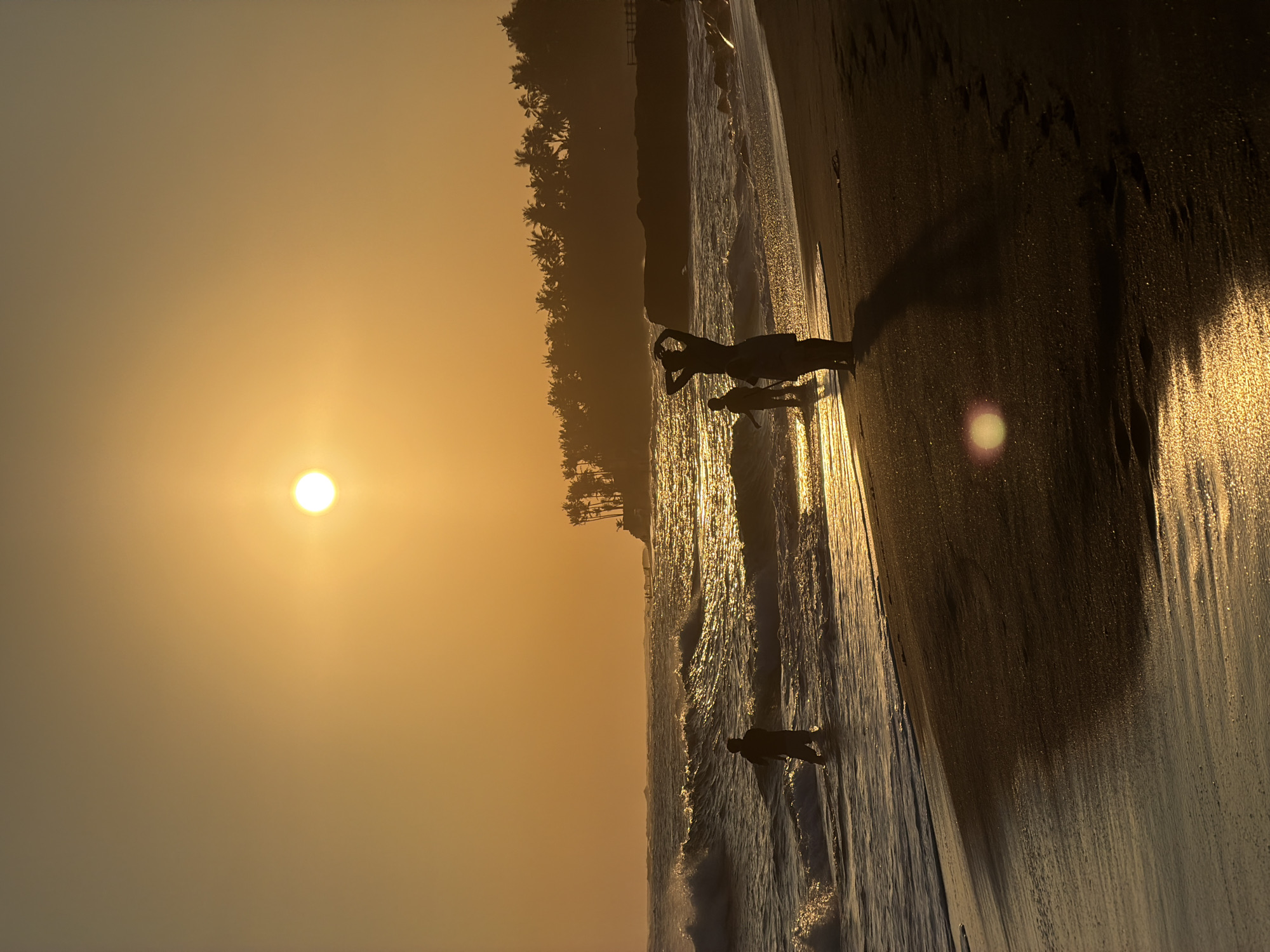 The image depicts a beach scene at sunset in Mengwi, Indonesia. The sun is low in the sky, casting a golden light across the scene. Silhouetted palm trees line the coast. Several individuals are seen on the beach, with one person in the foreground appearing to be swimming or floating in the water. A pair of figures stand further back, one with their arms raised. The beach is wet, reflecting the sunlight and the silhouettes of the people. Waves are visible near the shore. The overall atmosphere suggests a tranquil evening at the beach.