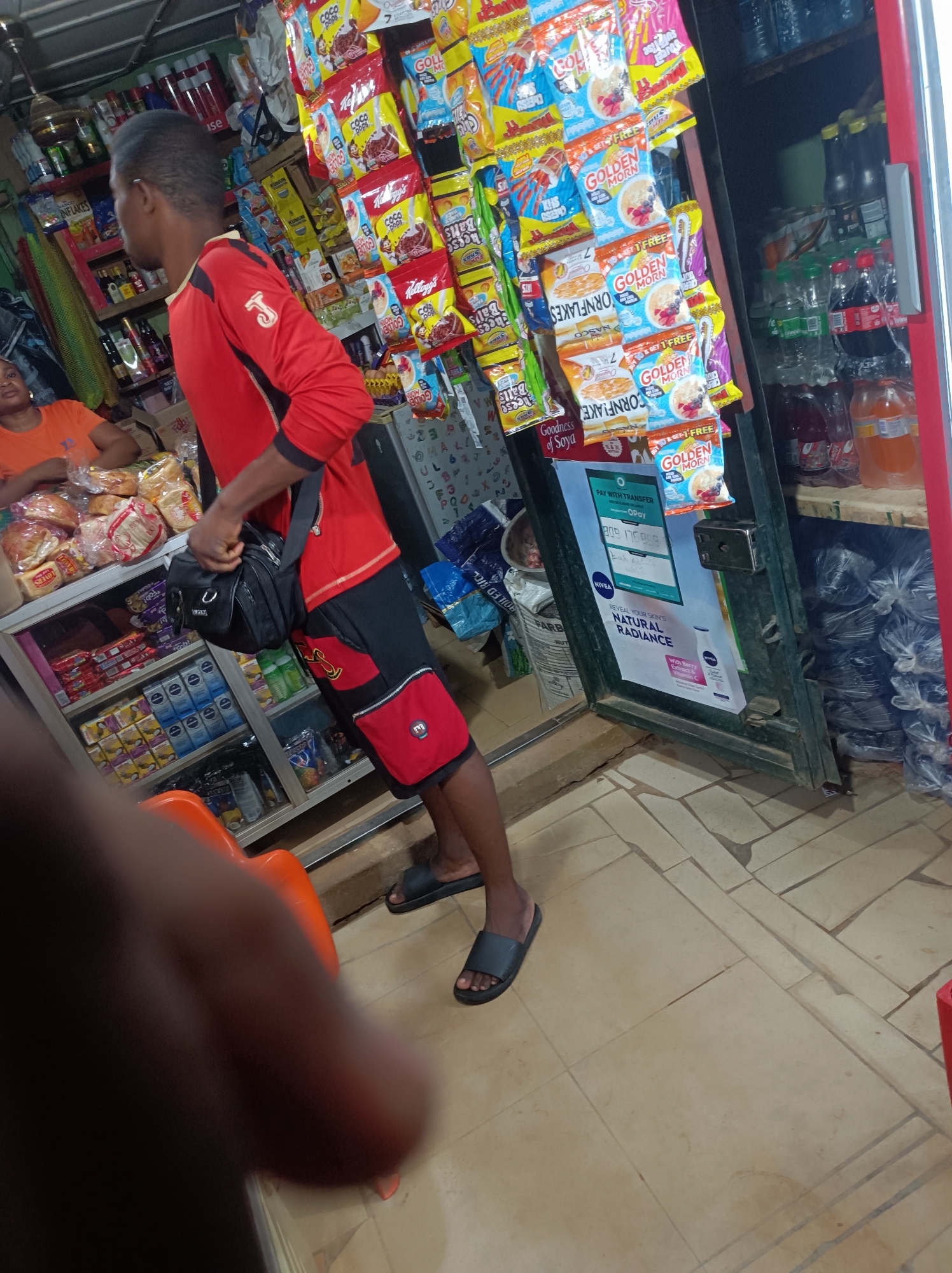 An indoor scene captures the bustling interior of a small shop, likely a minimart or provision store in Narayi, Nigeria. The primary subject is a man, seen from the side and back, standing near the center of the frame. He wears a red long-sleeved shirt with a black panel on the sleeves and a yellow "J" emblem, black shorts with red pockets, and black slide sandals. A black shoulder bag is slung across his body.
The shop is densely stocked with various consumer goods. To the man's right, numerous snack packets and cereal pouches are hung on display, including "GOLDEN MORN," "CORNFLAKE," "Coco Pops," "CHEESY Balls," and "Kellogg's," some advertising "GET 1 FREE." Next to this display, a refrigerator or cabinet is filled with bottled water and soft drinks. On the left side of the frame, another person, possibly a shop attendant, is partially visible sitting behind a glass display counter filled with packaged items like milk or powdered drinks, and loaves of bread in plastic bags. More shelves stocked with goods line the wall behind them.
A dark-colored door or door frame stands open to the right, featuring a "NIVEA" advertisement that reads "REVEAL YOUR SKIN'S NATURAL RADIANCE" and a payment sign for "OPay" with "PAY WITH TRANSFER" and the number "170959." Black plastic-wrapped items are stacked on the floor near the door, and a white sack labeled "BOILED RIC" and "PARB" is visible further back. The floor is tiled in a light brown pattern, and a red plastic chair is partially visible in the foreground. The scene depicts a typical daily activity in a local retail establishment.